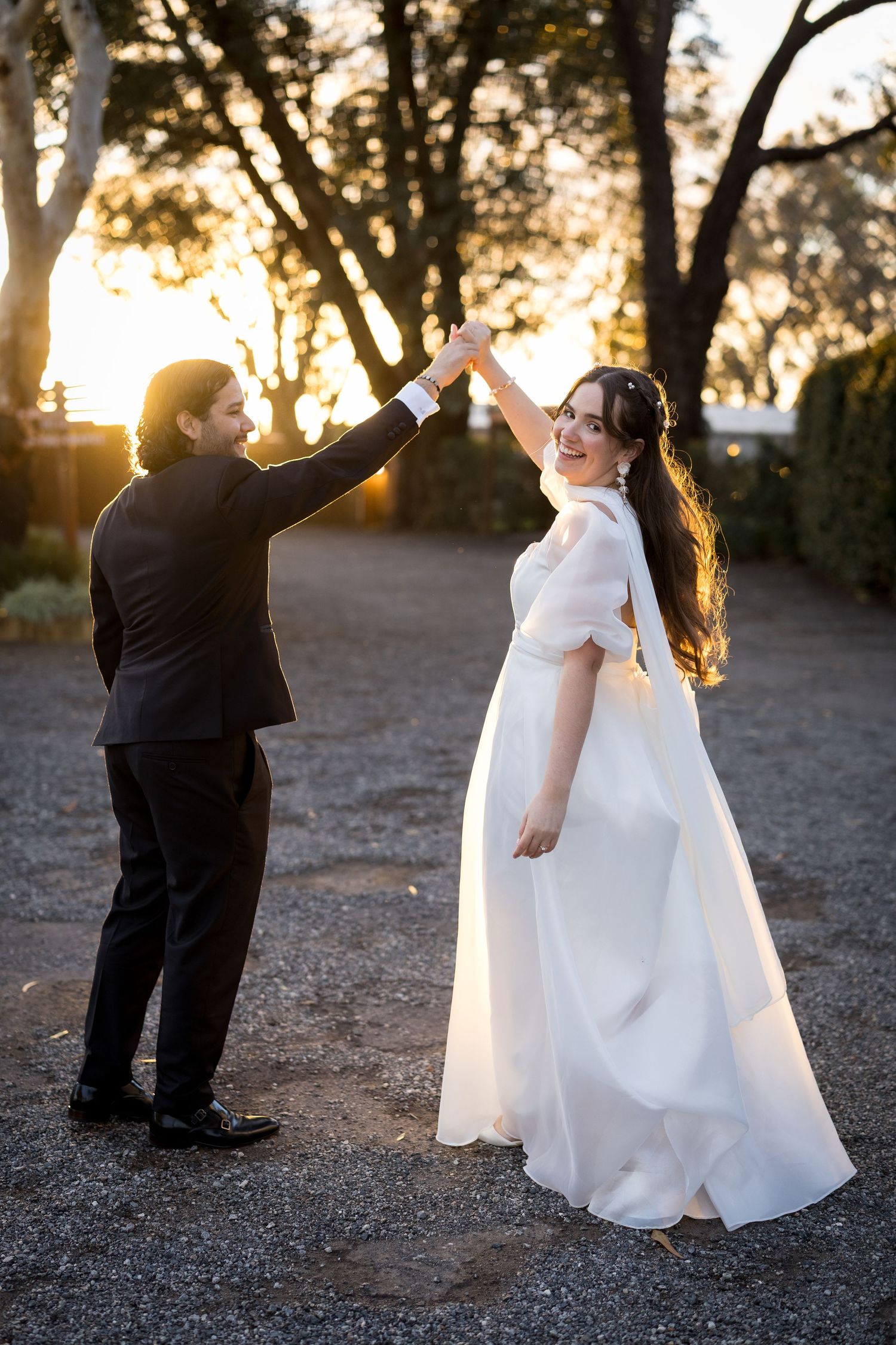 Bride in white gown twirls as groom in black suit holds her hand at golden hour sunset outdoors.