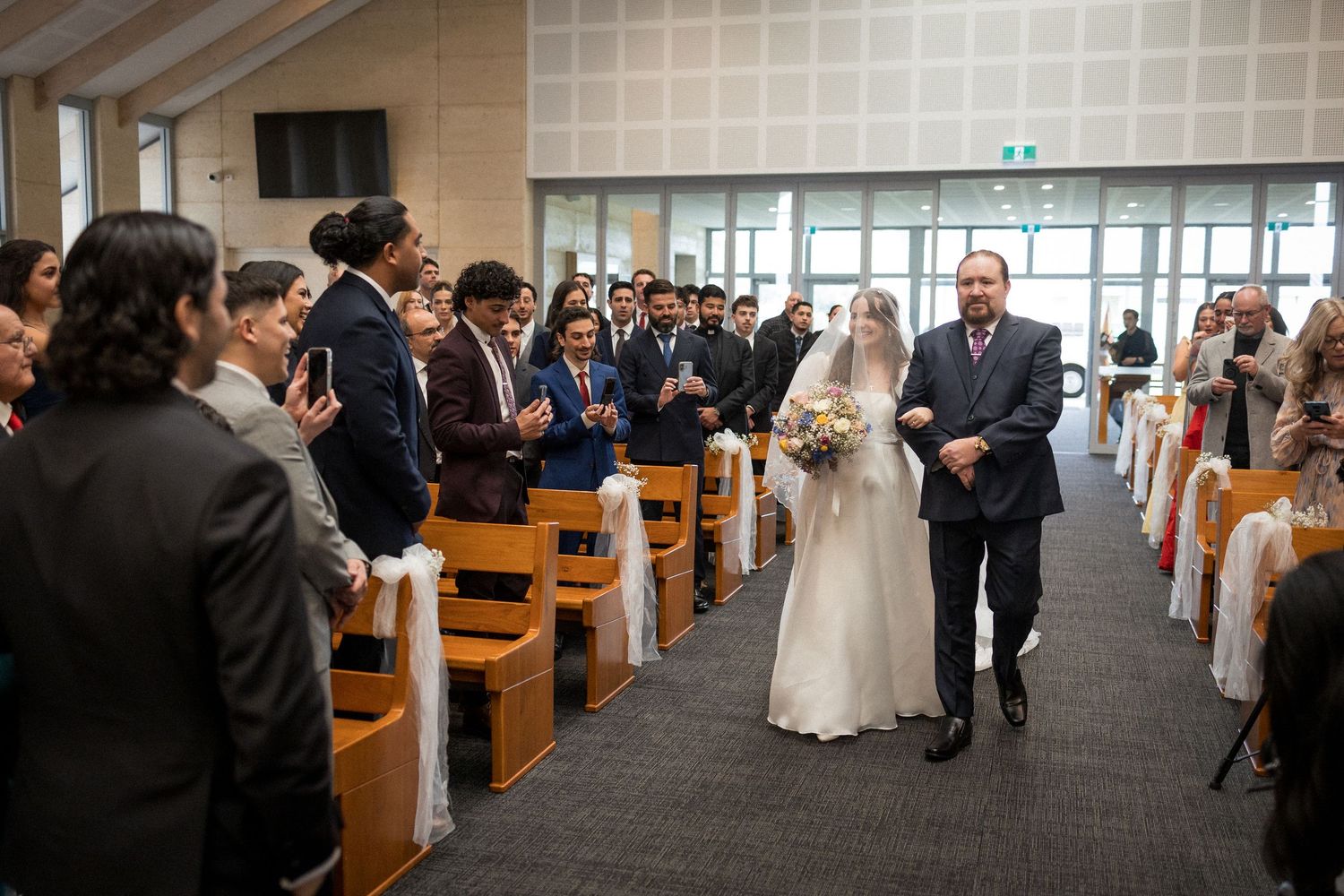 Bride walks down church aisle with escort as wedding guests stand and applaud the ceremony.