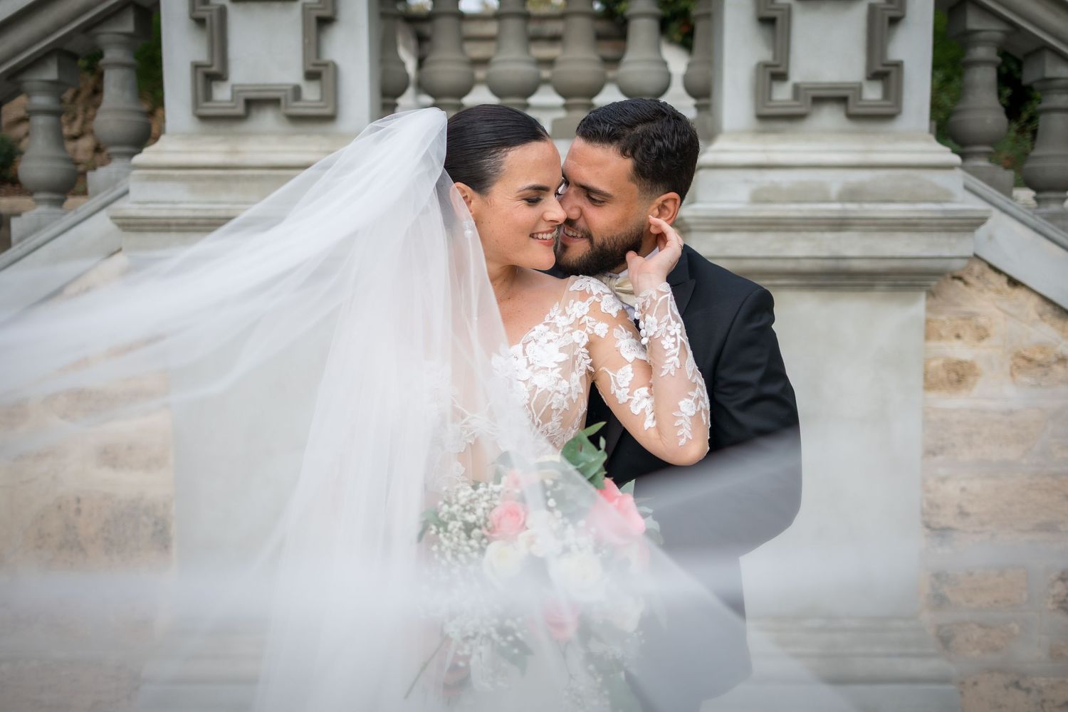 Bride and groom share a romantic moment on stone steps as her lace veil billows around them beautifully.