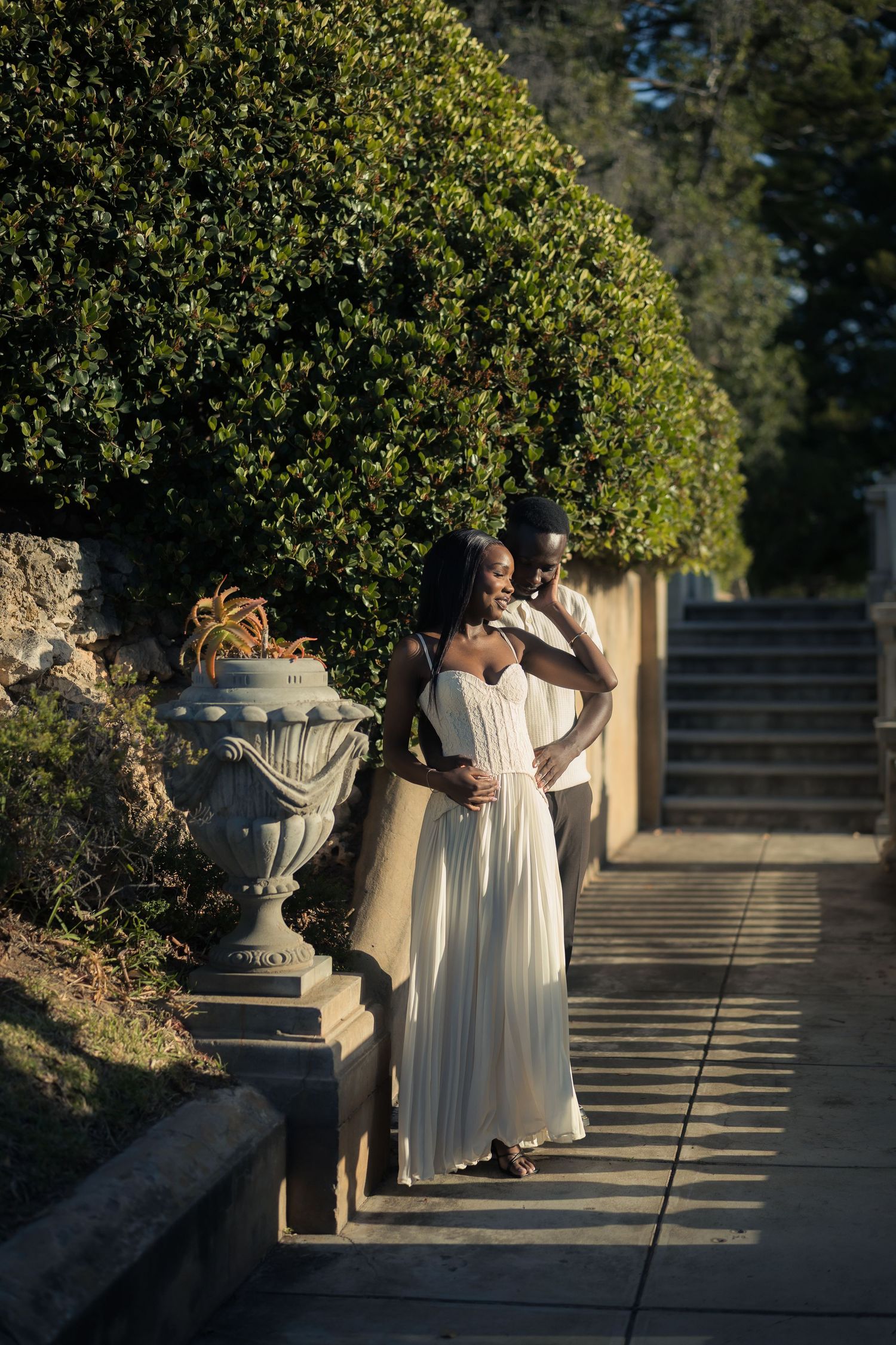 Couple embracing in golden sunlight beside an ornate stone urn and green hedge on a garden path, the woman in a flowing white dress.