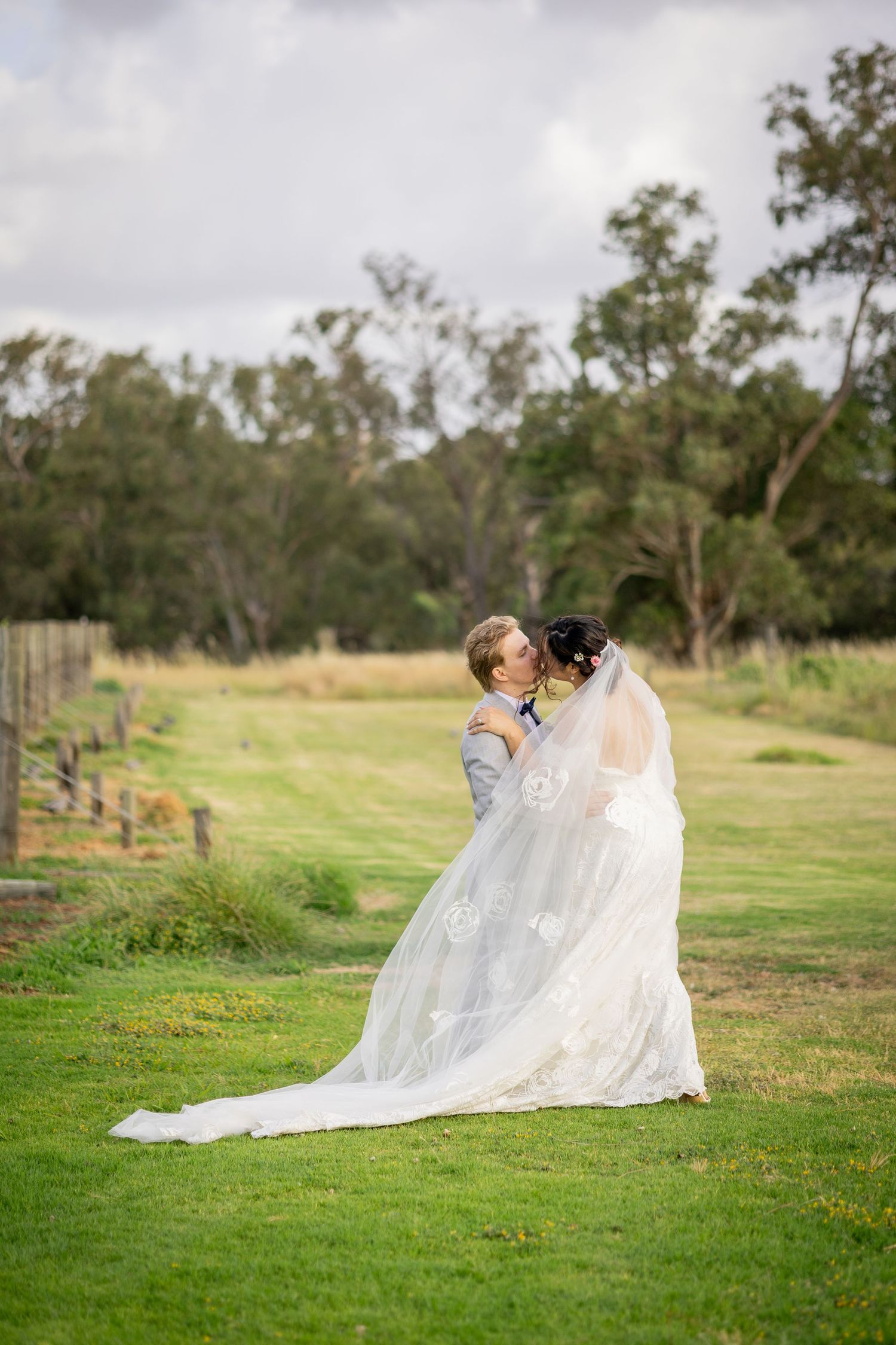 Bride and groom share a romantic kiss on lush green lawn with flowing lace wedding veil and forest backdrop.