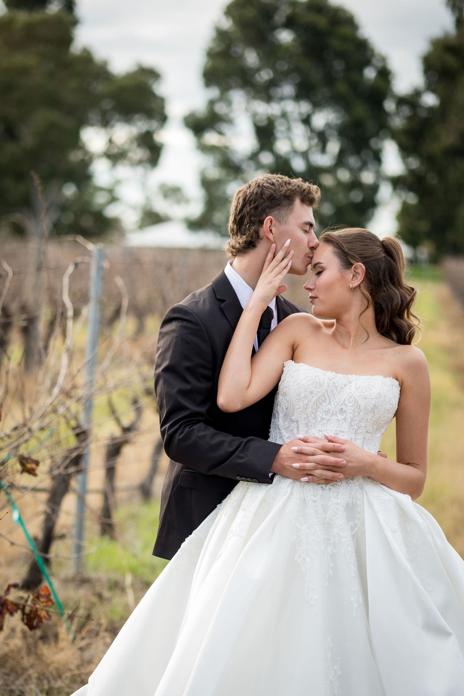 Bride and groom sharing a tender kiss in a vineyard, bride in strapless white gown, groom in dark suit.