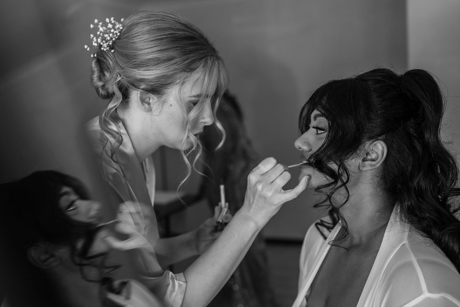 Bride applying makeup to bridesmaid in elegant black and white wedding preparation photo.