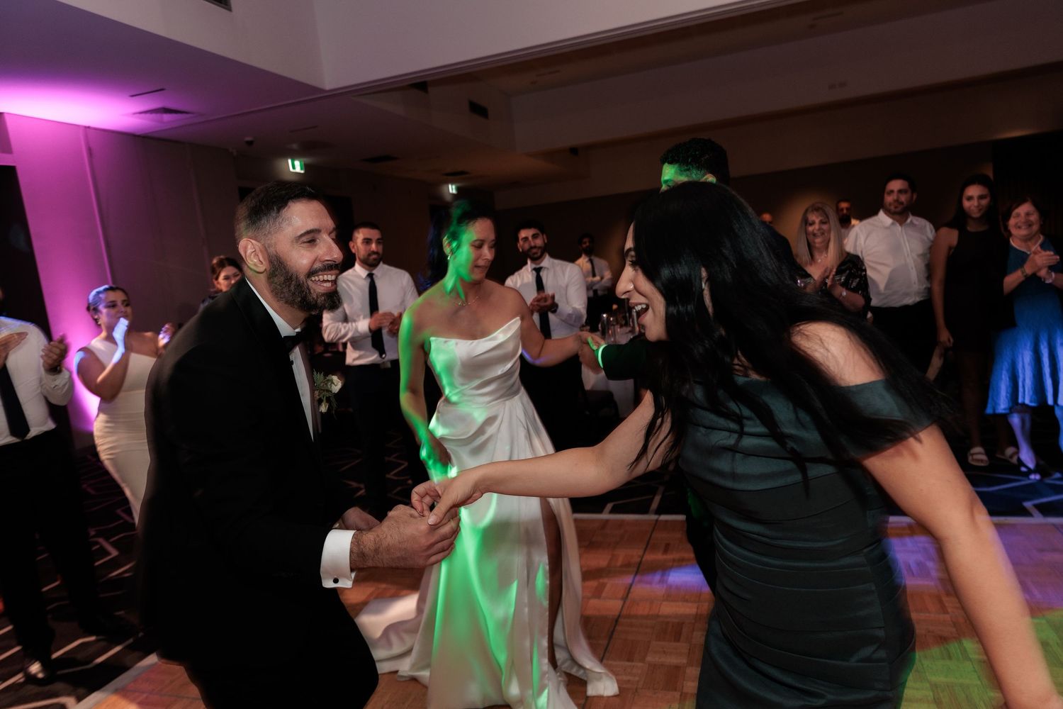 Bride and groom dance joyfully with guests on the wedding reception dance floor under green and pink lights.