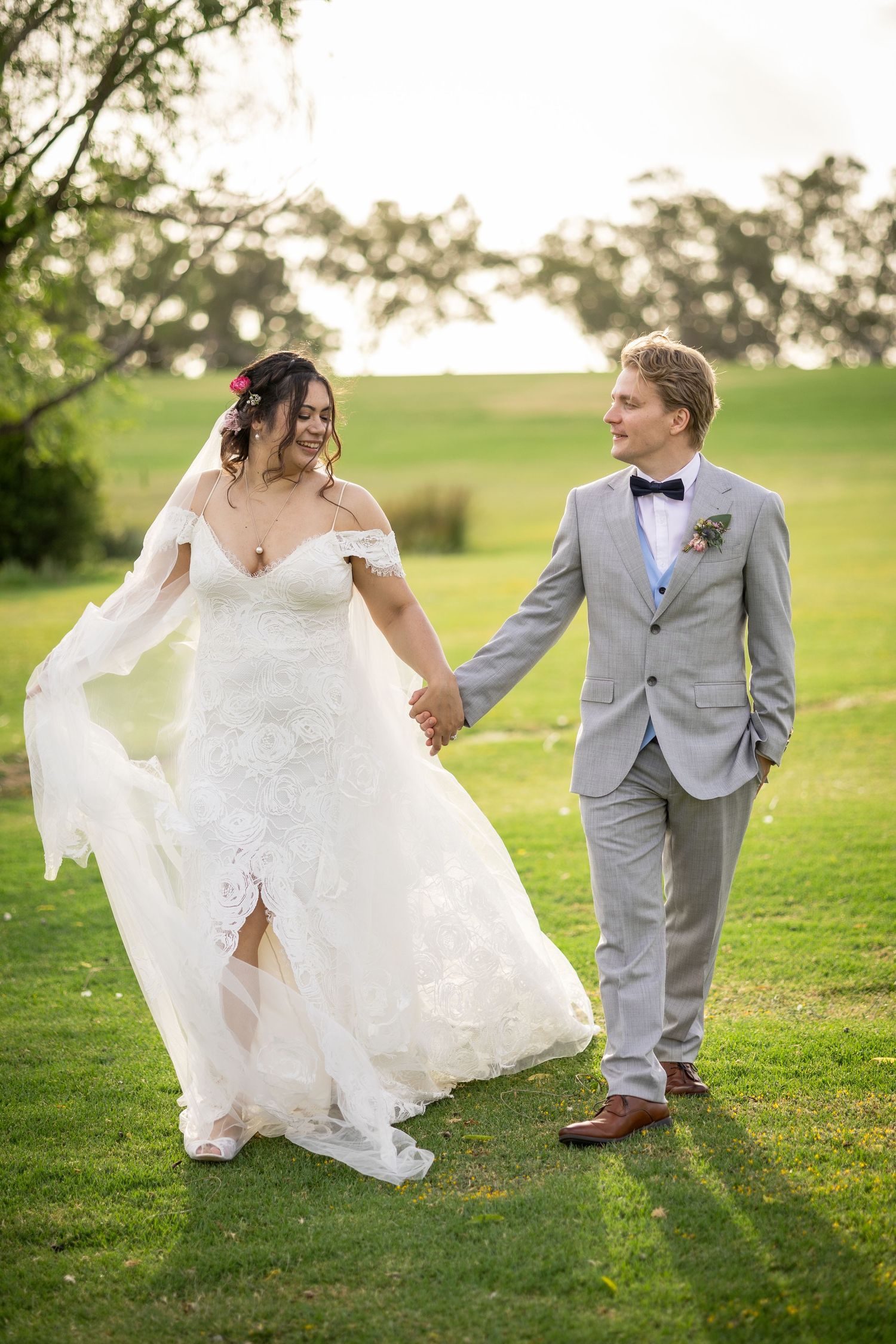 Bride and groom walk hand-in-hand on green lawn, she in lace gown, he in gray suit with boutonniere.