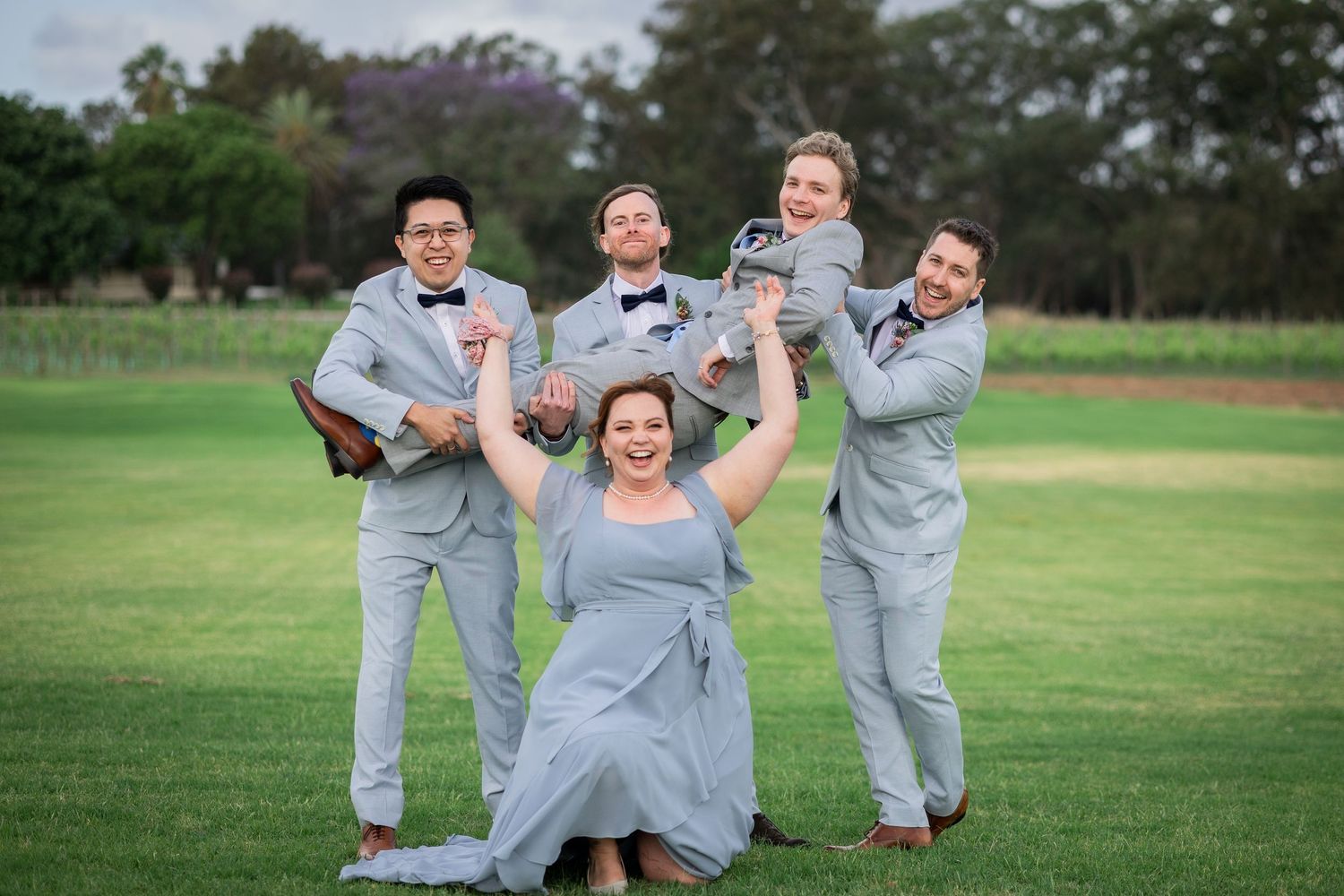 Wedding party posing playfully on green grass, groomsmen in grey suits lifting bride in blue dress.