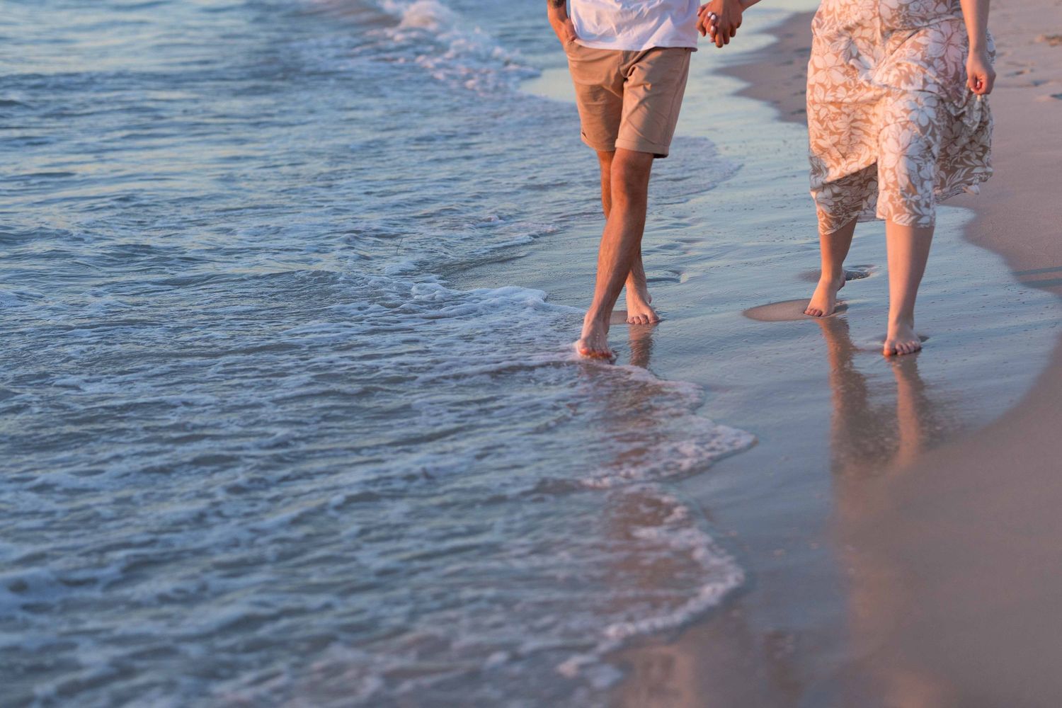 Couple walking barefoot on a sandy beach at sunset, waves gently washing over their feet.