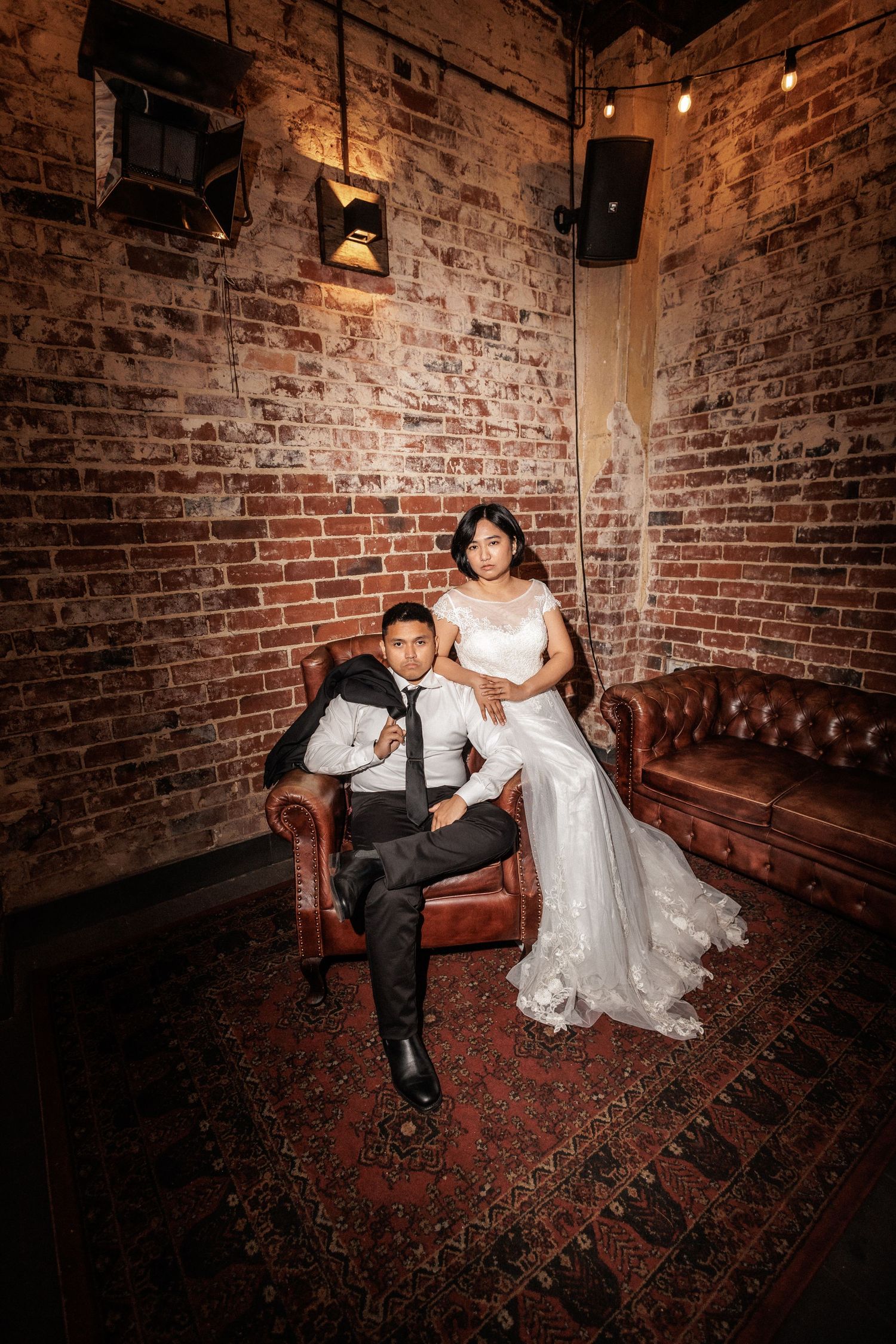 Bride in white lace gown sits on leather sofa beside suited groom against rustic brick wall in moody venue.