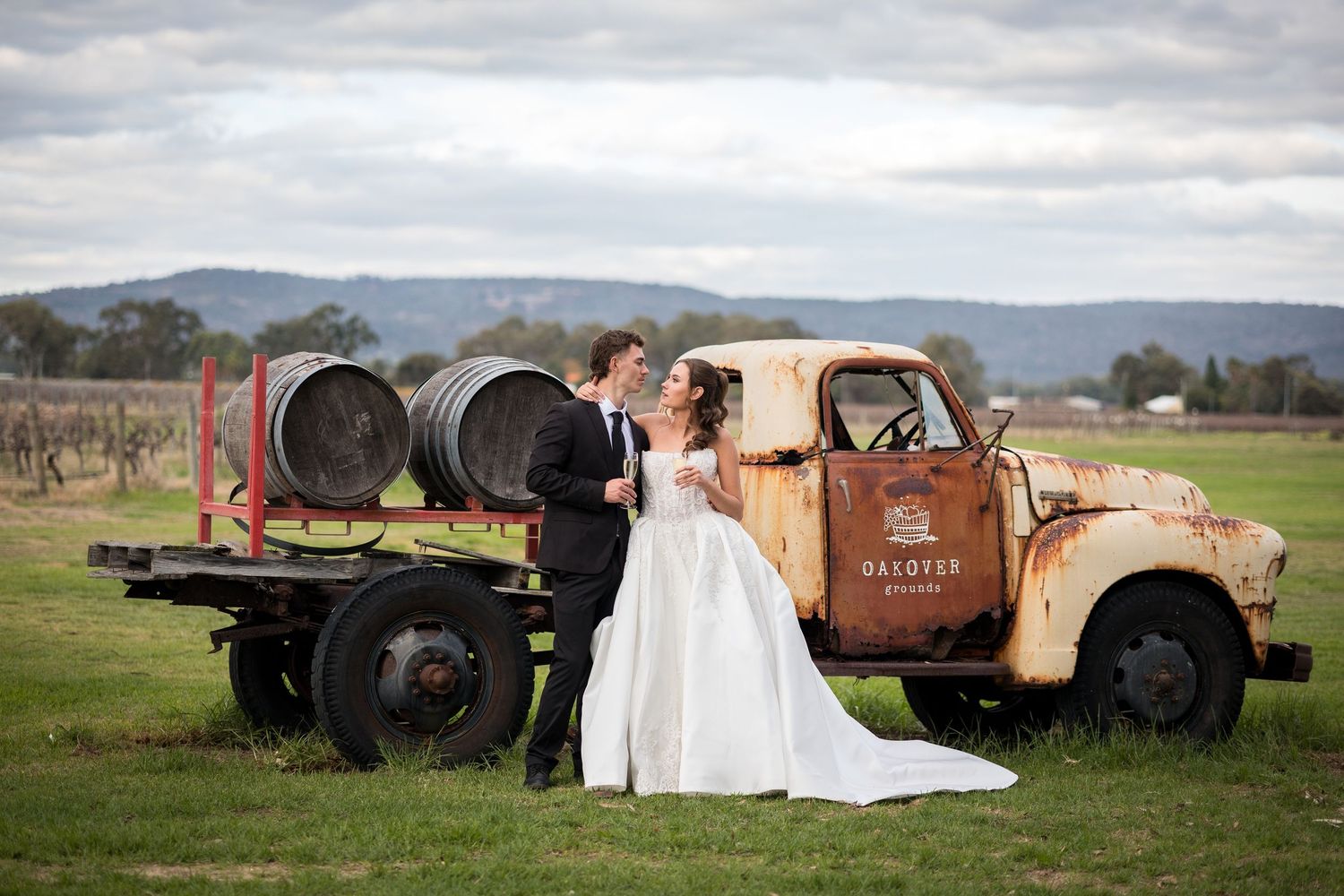 Bride and groom posing beside a rustic vintage truck with wine barrels at a winery vineyard wedding venue.