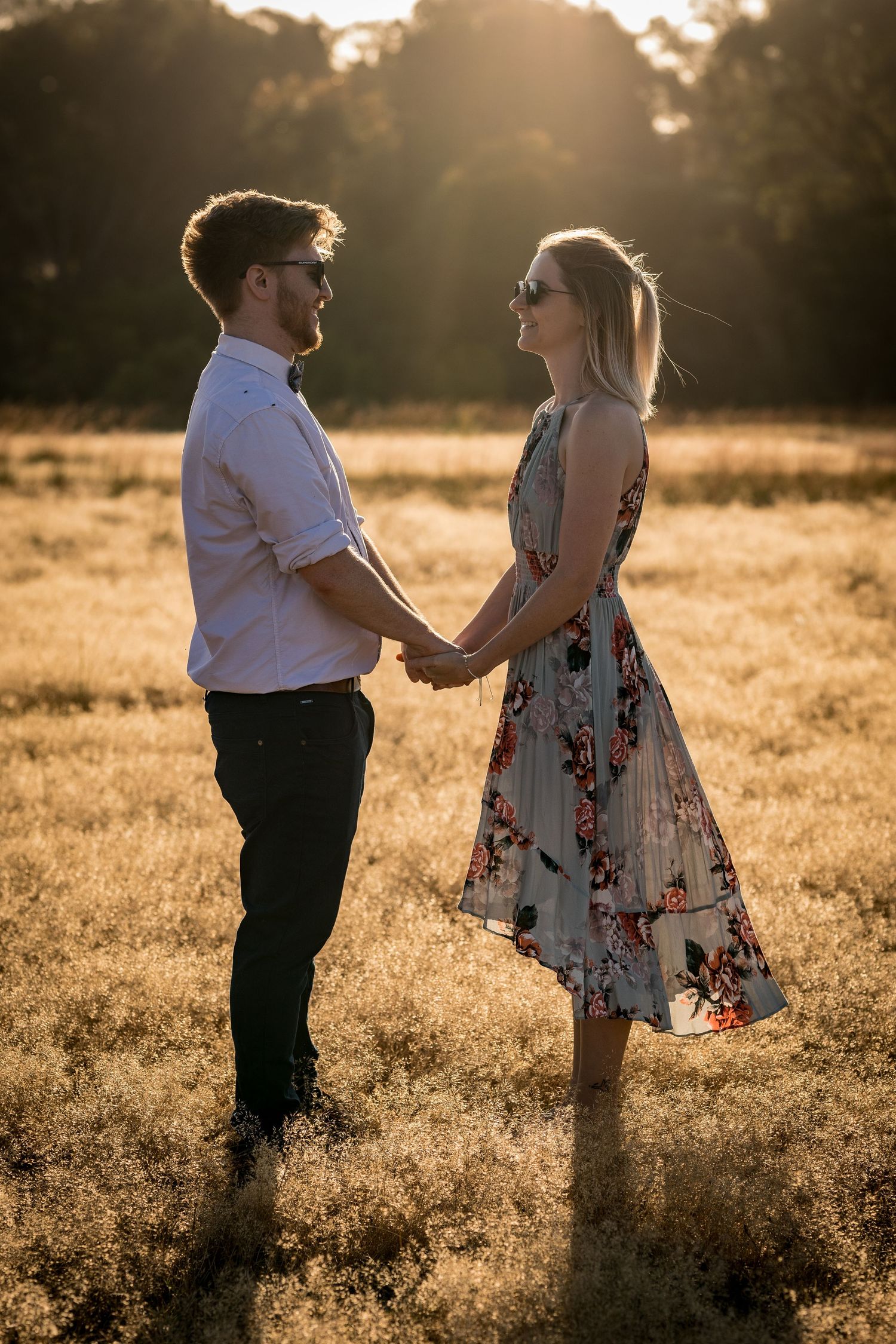 A couple holds hands in a golden sunlit field, the woman in a floral dress and the man in a white shirt.