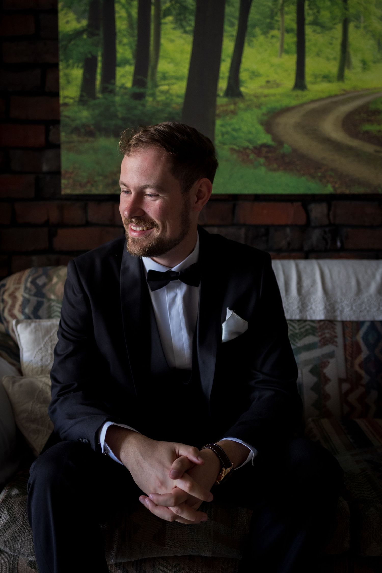 Smiling groom in black tuxedo with bow tie sits relaxed with clasped hands before a lush forest painting backdrop.