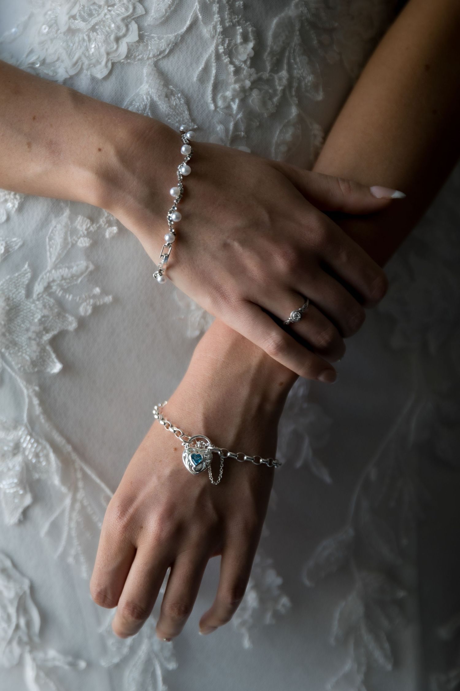 Bride wearing two silver bracelets, one with crystals and one with a blue heart charm, crossed hands over lace wedding dress.