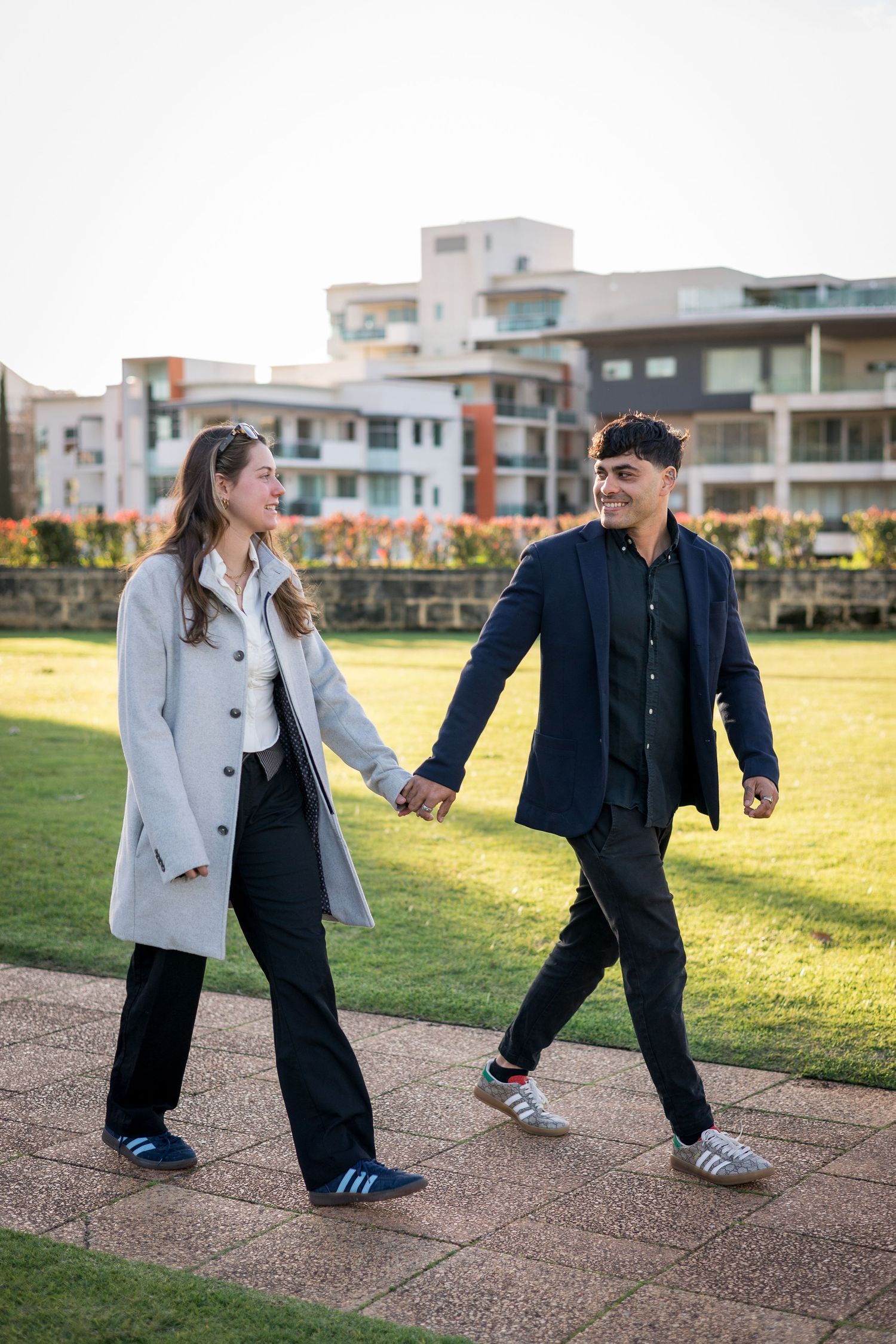 Playful couple dancing on a path with modern buildings and green lawn in the background.