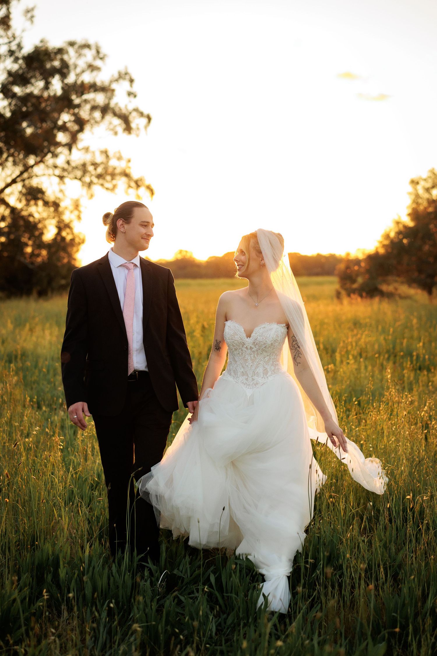 A bride and groom walk hand-in-hand through a sunlit green field at golden hour, smiling at each other.