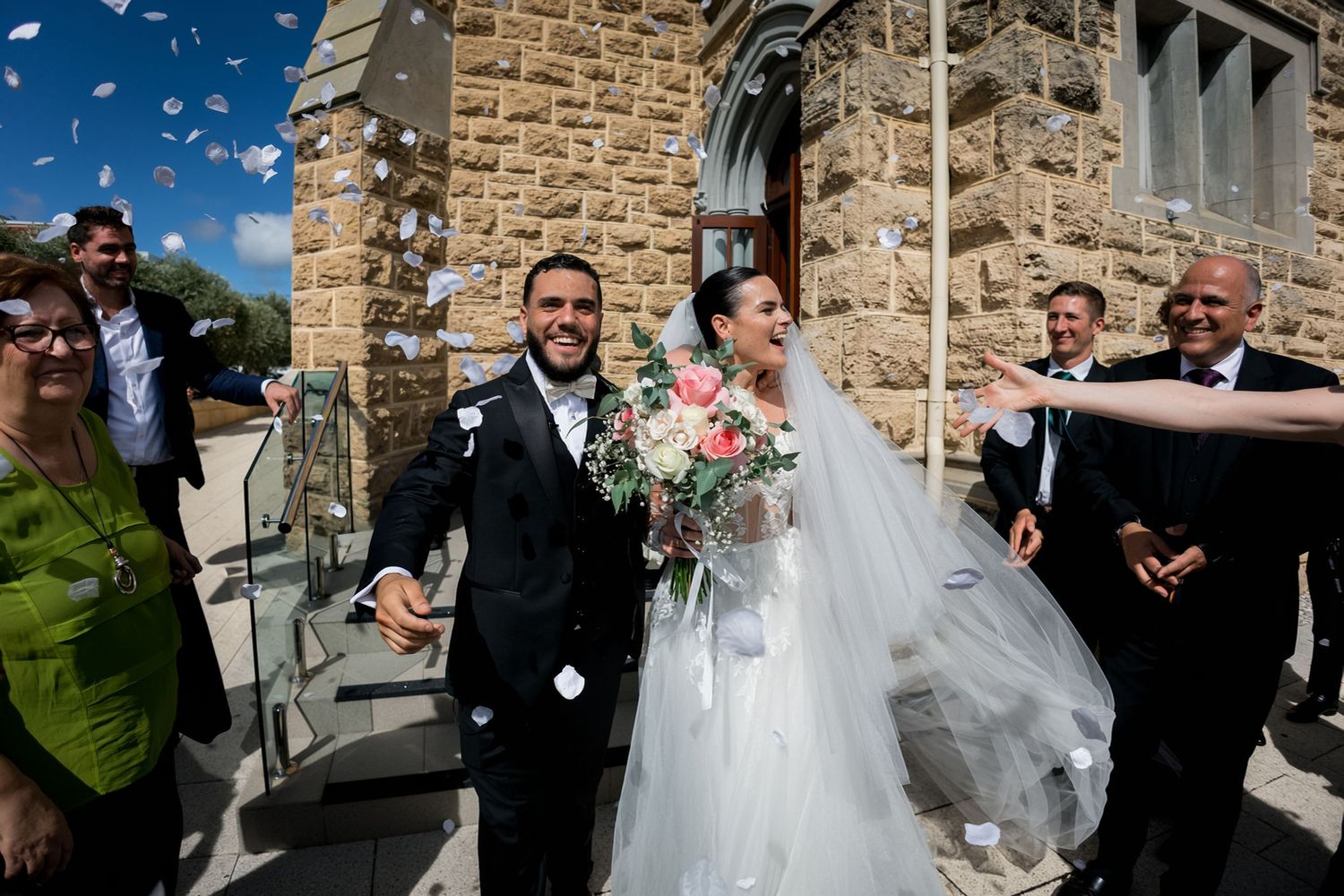 Bride and groom exit stone church as guests throw confetti, bride holding pink bouquet in white gown.