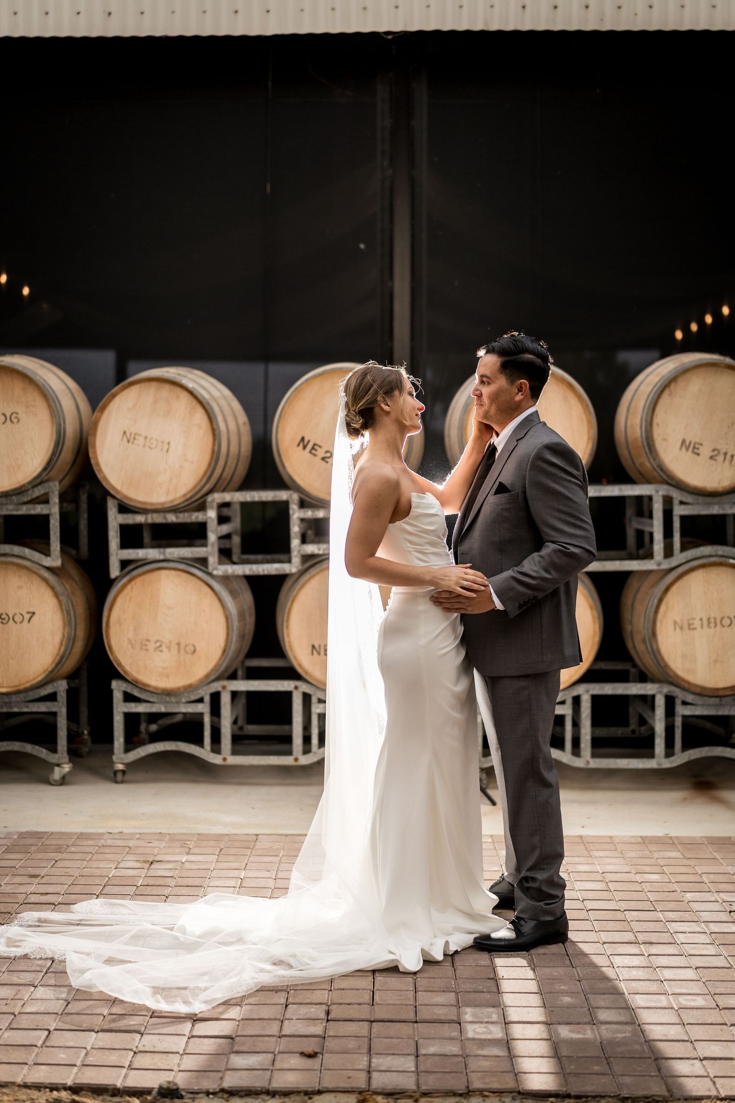 Bride and groom sharing a tender moment in front of stacked oak wine barrels at a winery.