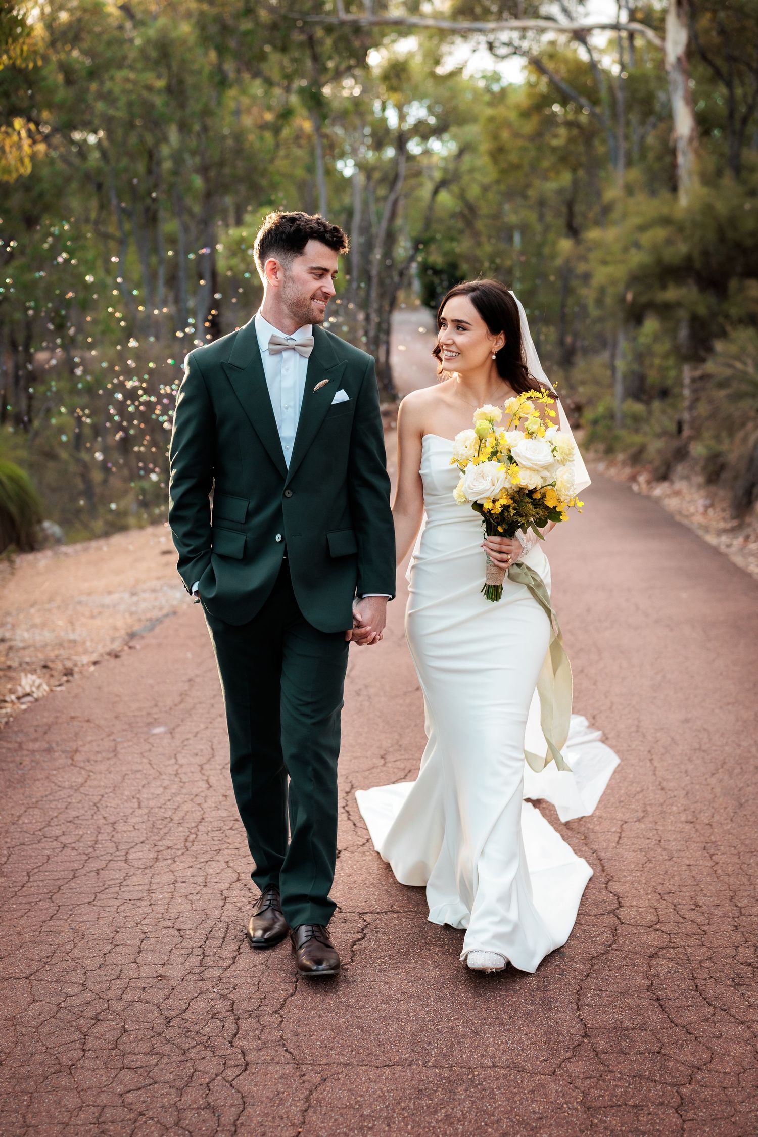 Bride and groom walk hand-in-hand on a red path, she holds yellow bouquet, he wears dark green suit.
