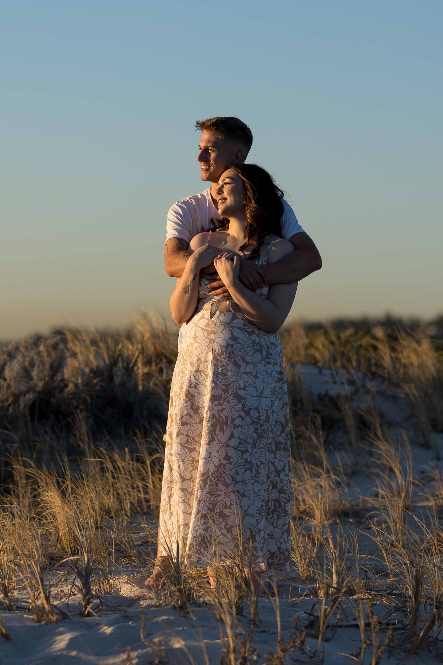 Couple embracing on sandy dunes at sunset, woman in floral dress leaning back into partner.