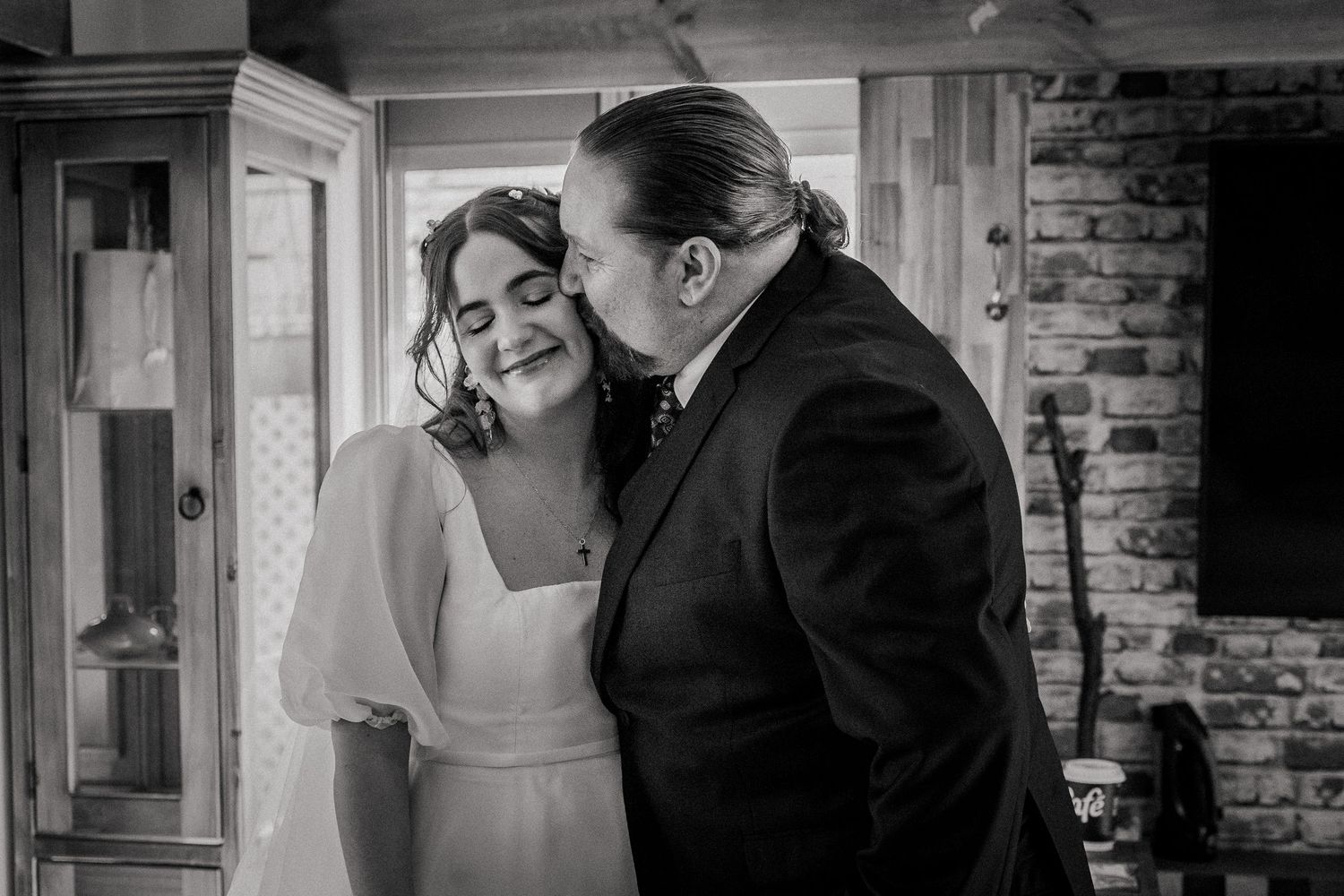 Black and white wedding photo of bride and groom sharing a tender moment, groom kissing bride's cheek indoors.