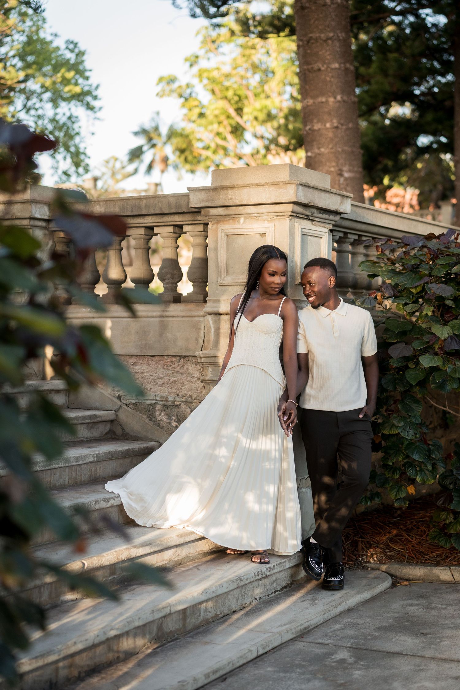 Couple in coordinated cream and black outfits smiling together on stone garden steps surrounded by lush greenery at golden hour.