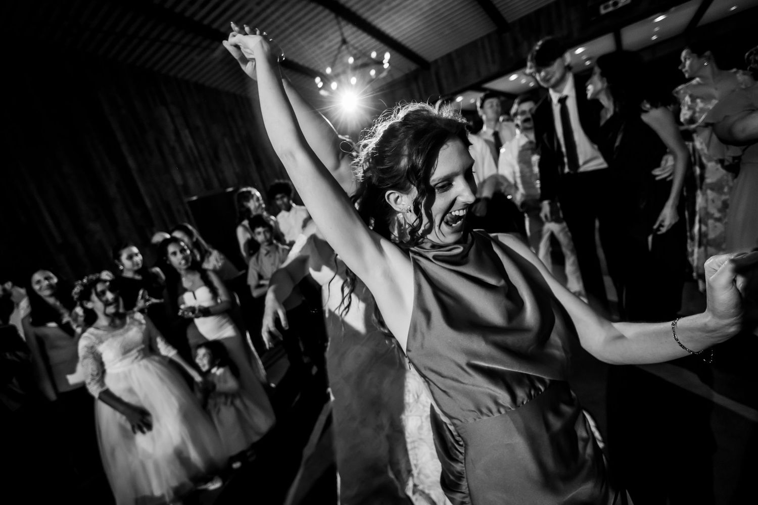Black and white photo of a woman dancing joyfully with arms raised at a lively wedding reception celebration.