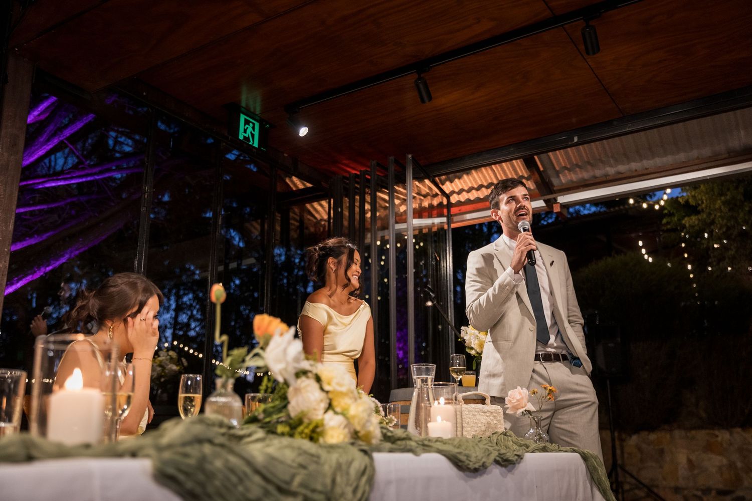 Groom gives speech at elegantly decorated wedding reception table with floral centerpieces and candlelight.