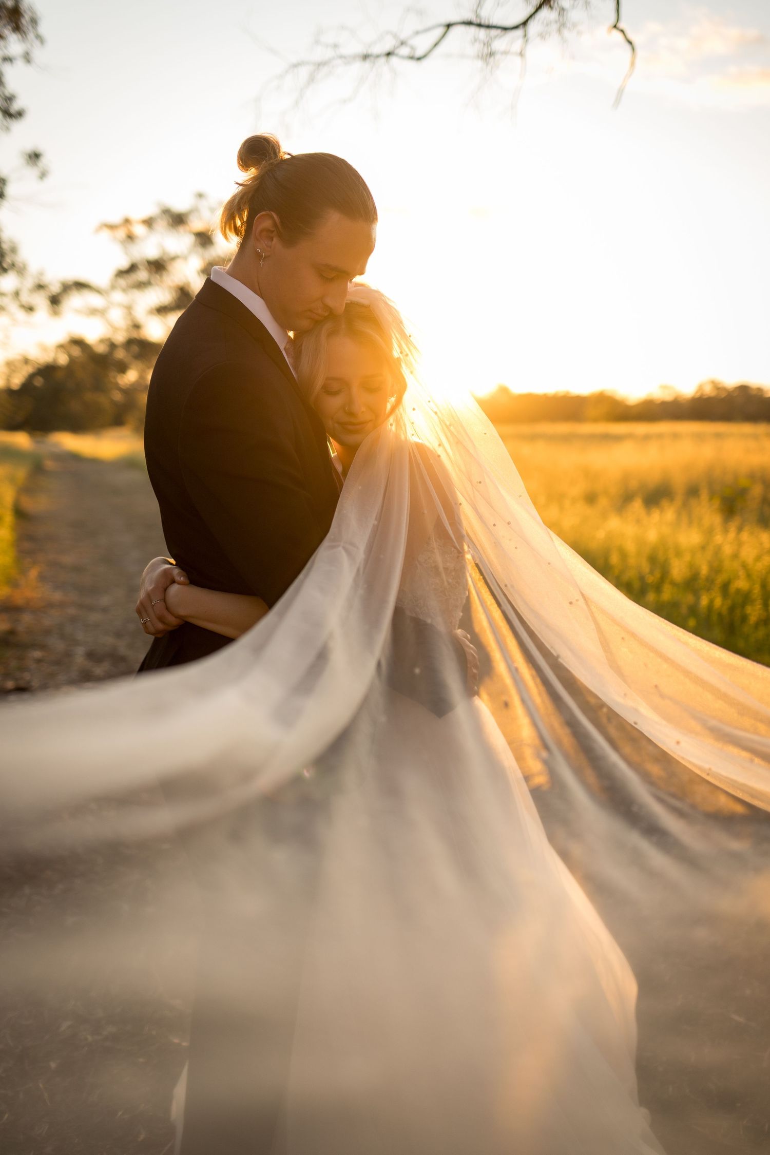 Couple embraces at golden sunset, bride's veil flowing in the breeze on a country path.