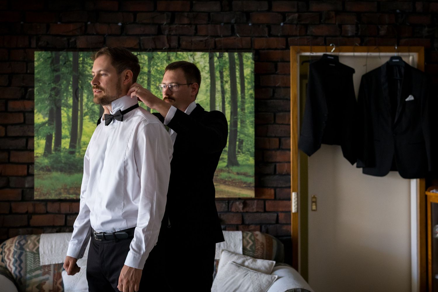 A man helps adjust another mans bow tie while getting ready in a rustic room with brick walls and hanging suits.
