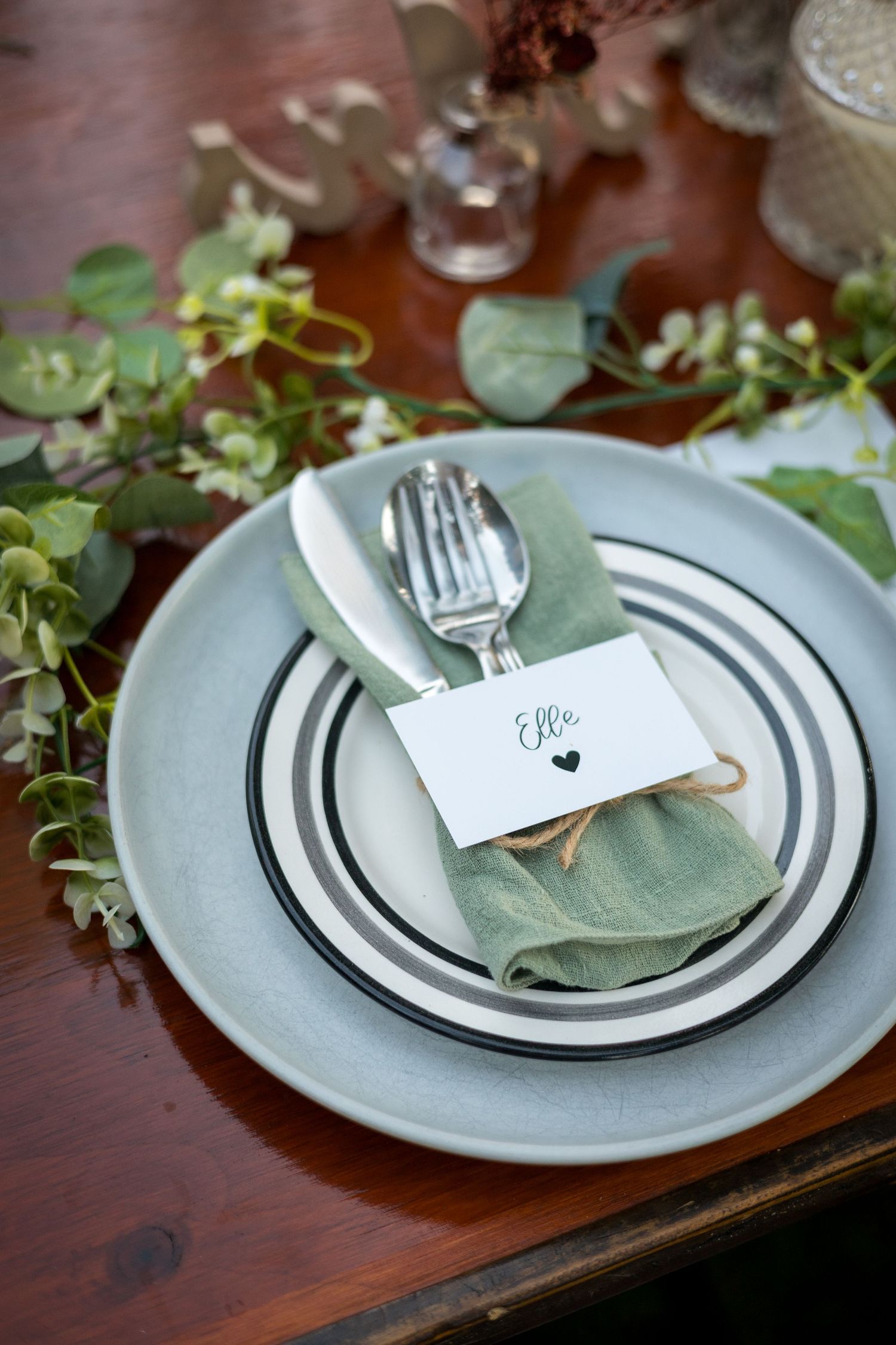Modern place setting with striped plates, sage napkin and name card on wooden table with greenery.