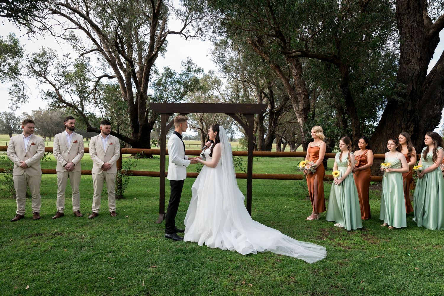 Wedding party poses for formal photos on a grassy lawn with trees in the background.