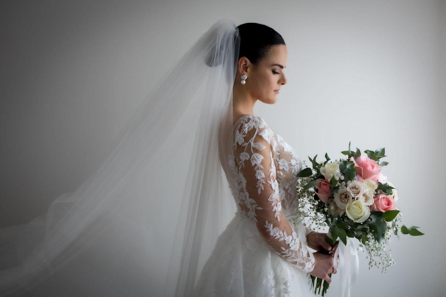 Elegant bride in lace long-sleeve wedding gown and veil holding pink and white bouquet, posing gracefully against white wall.