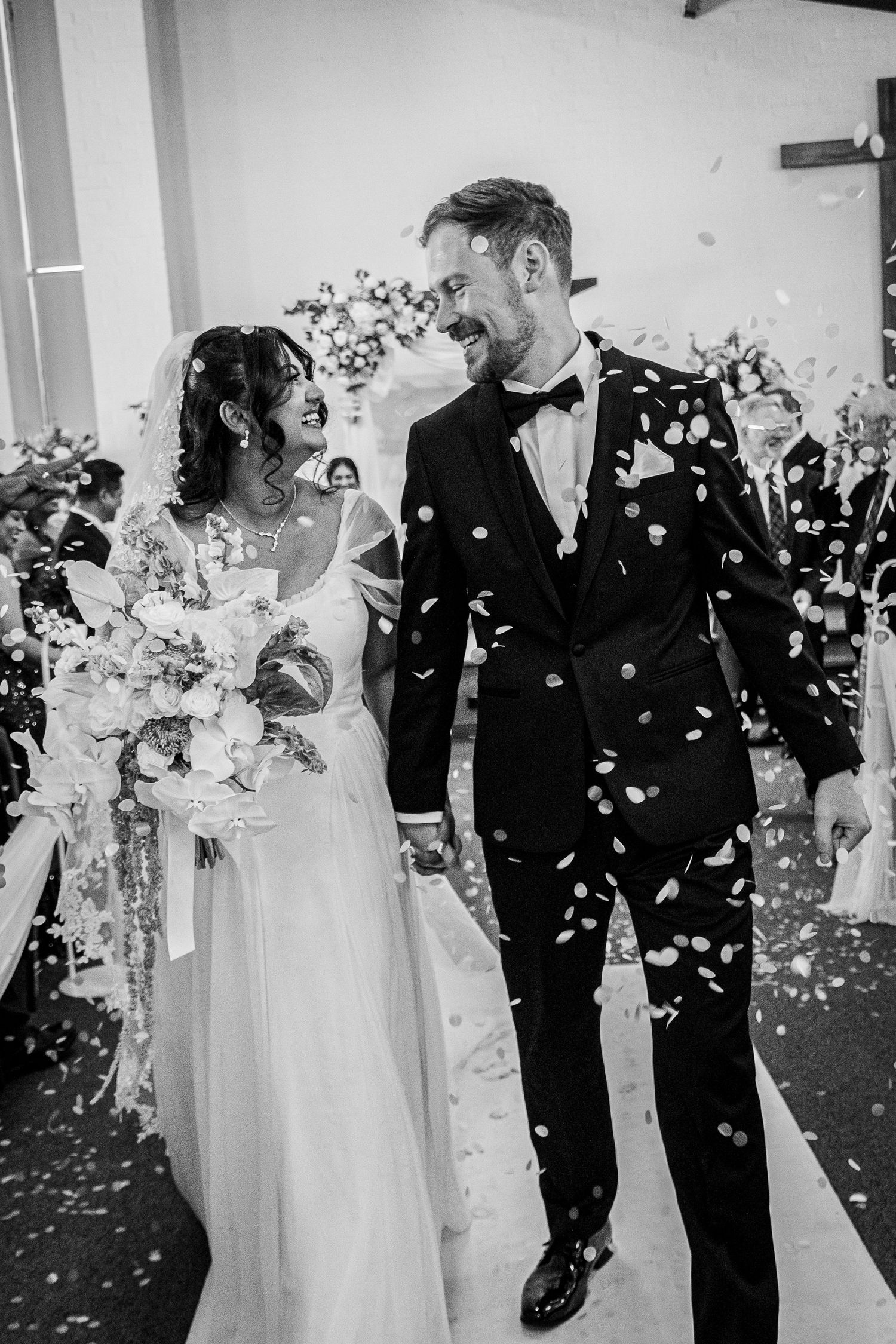 Black and white photo of a joyful bride and groom walking down the aisle as confetti falls around them.