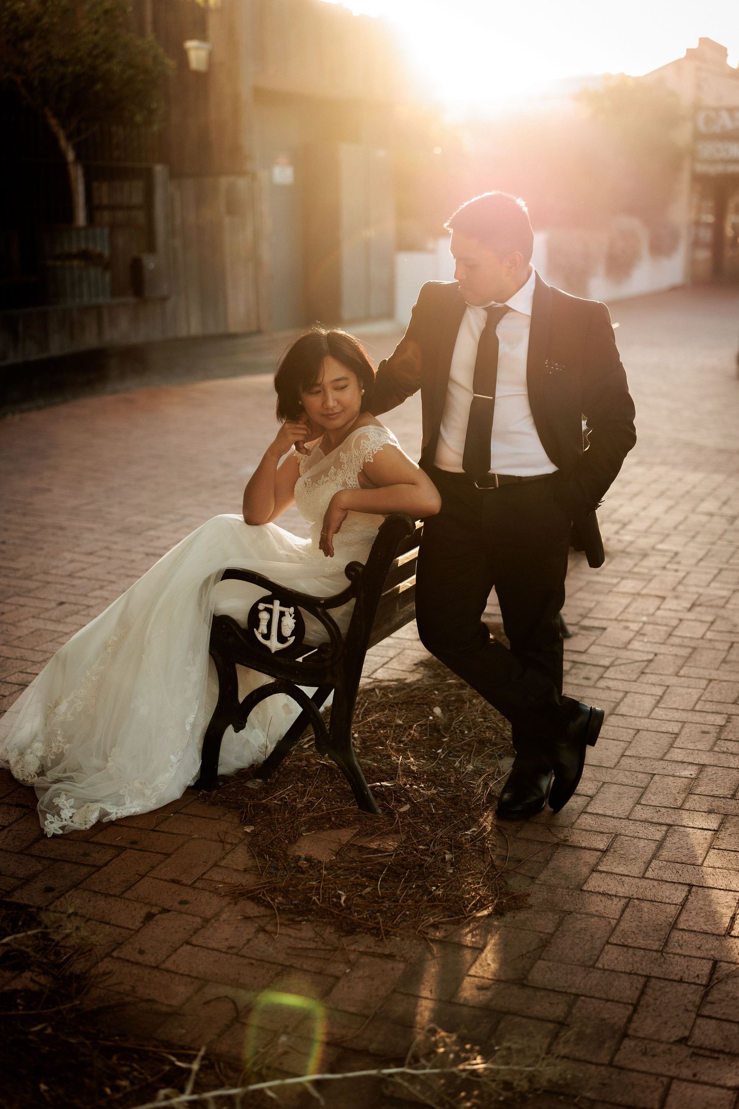 Bride in white gown sits on bench as groom in black suit leans close, bathed in golden sunset light on brick plaza.