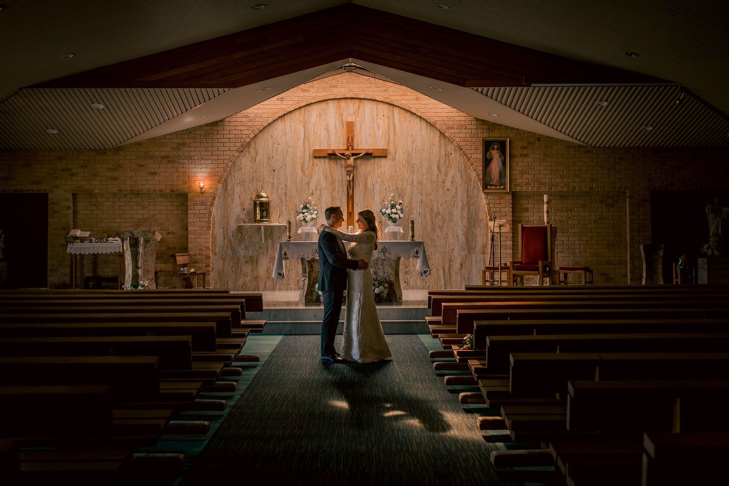 Bride and groom share a romantic embrace at the church altar, bathed in warm light with a crucifix backdrop.