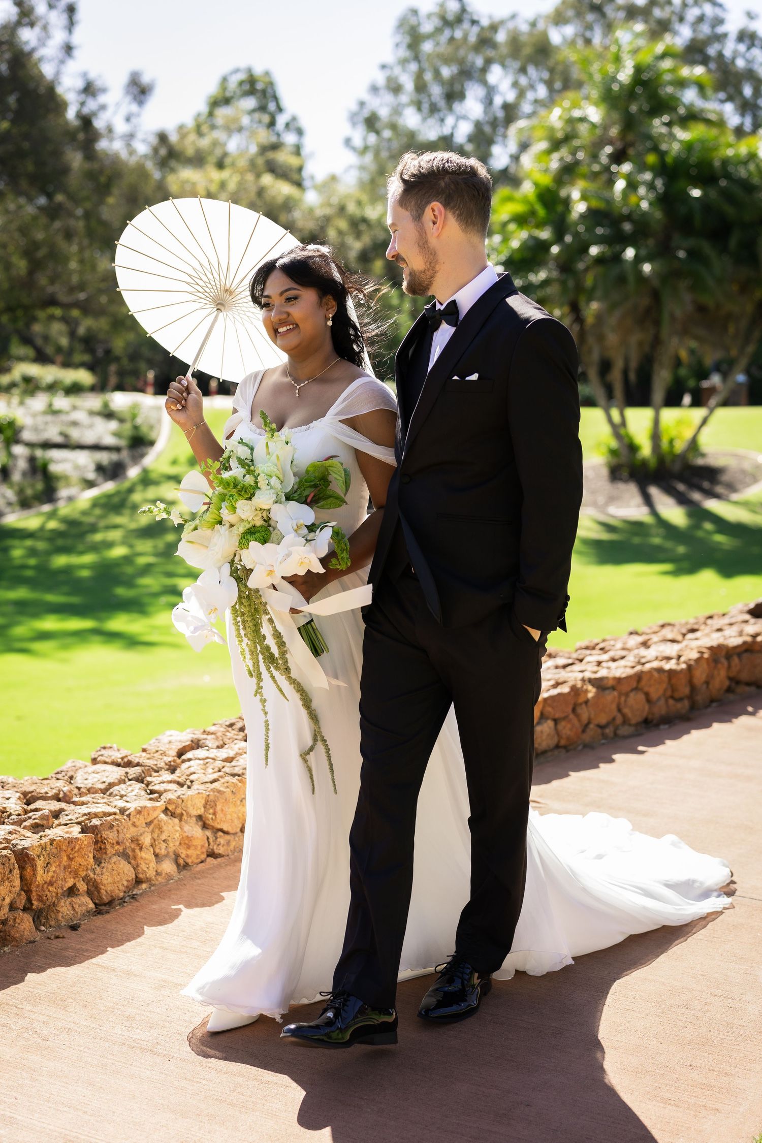 Bride holding white parasol and floral bouquet stands with groom in black tuxedo in sunny garden setting.