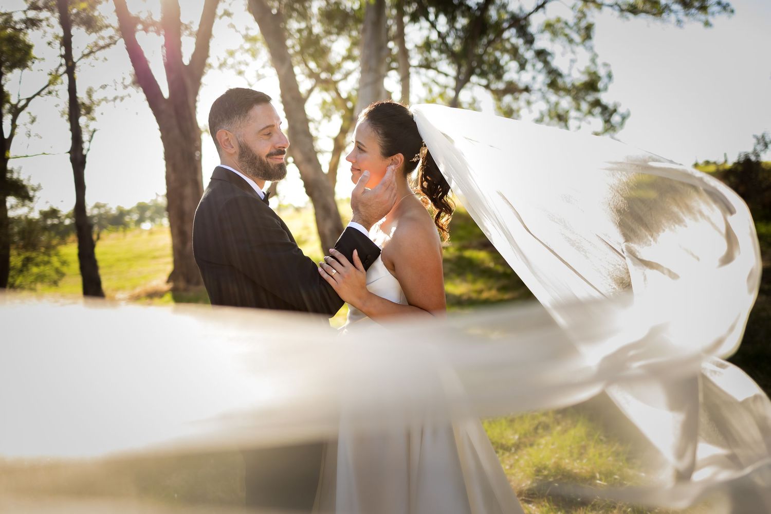 Bride and groom share romantic moment outdoors with wedding veil flowing in the breeze.