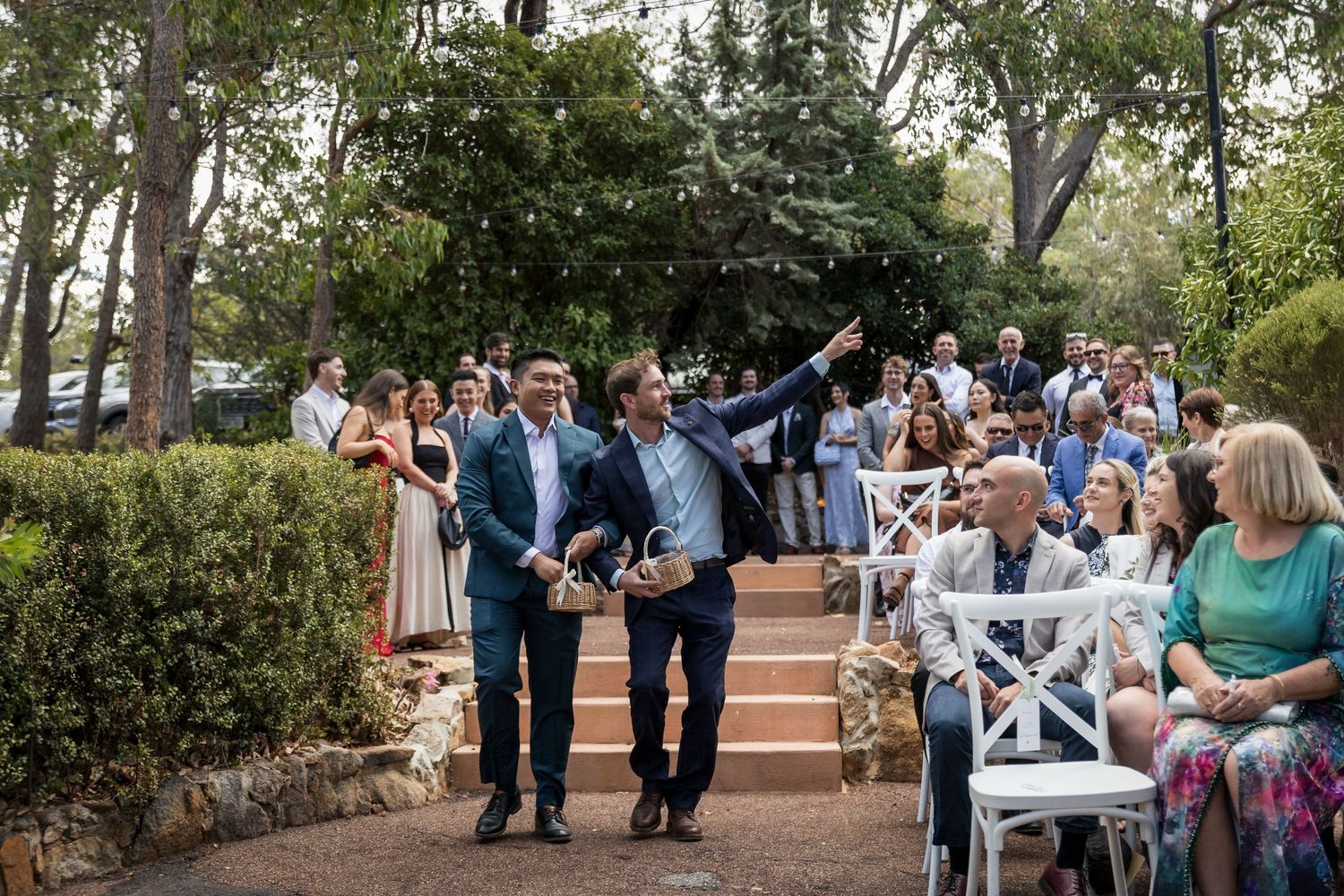 Two men scatter flower petals at an outdoor wedding ceremony while guests watch from white chairs in a garden setting.