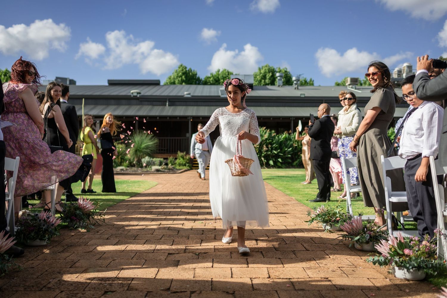 Flower girl in white dress walks down brick aisle at outdoor wedding ceremony with guests seated beside her.