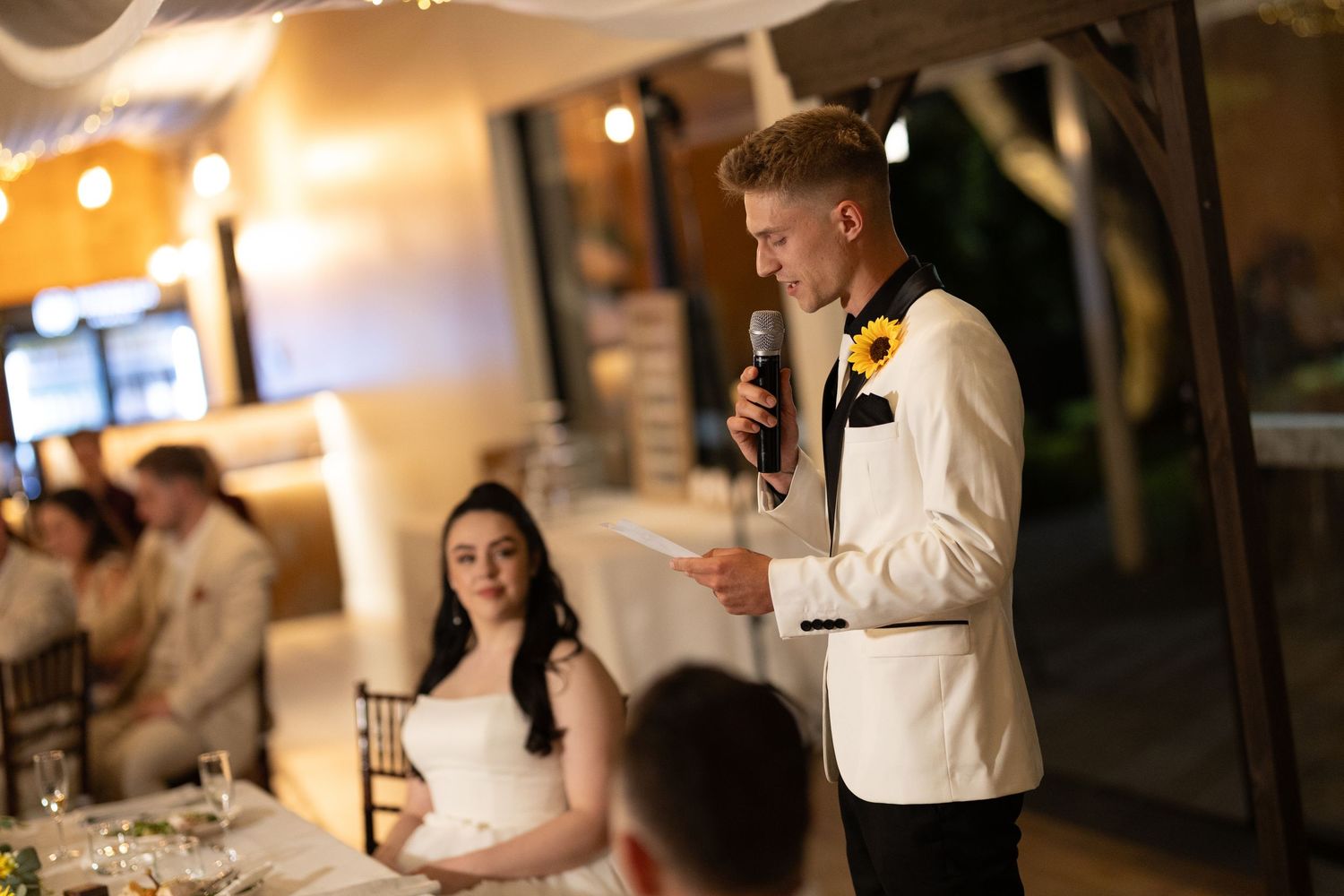Reception speech being delivered in a warmly lit dining room during wedding celebrations.