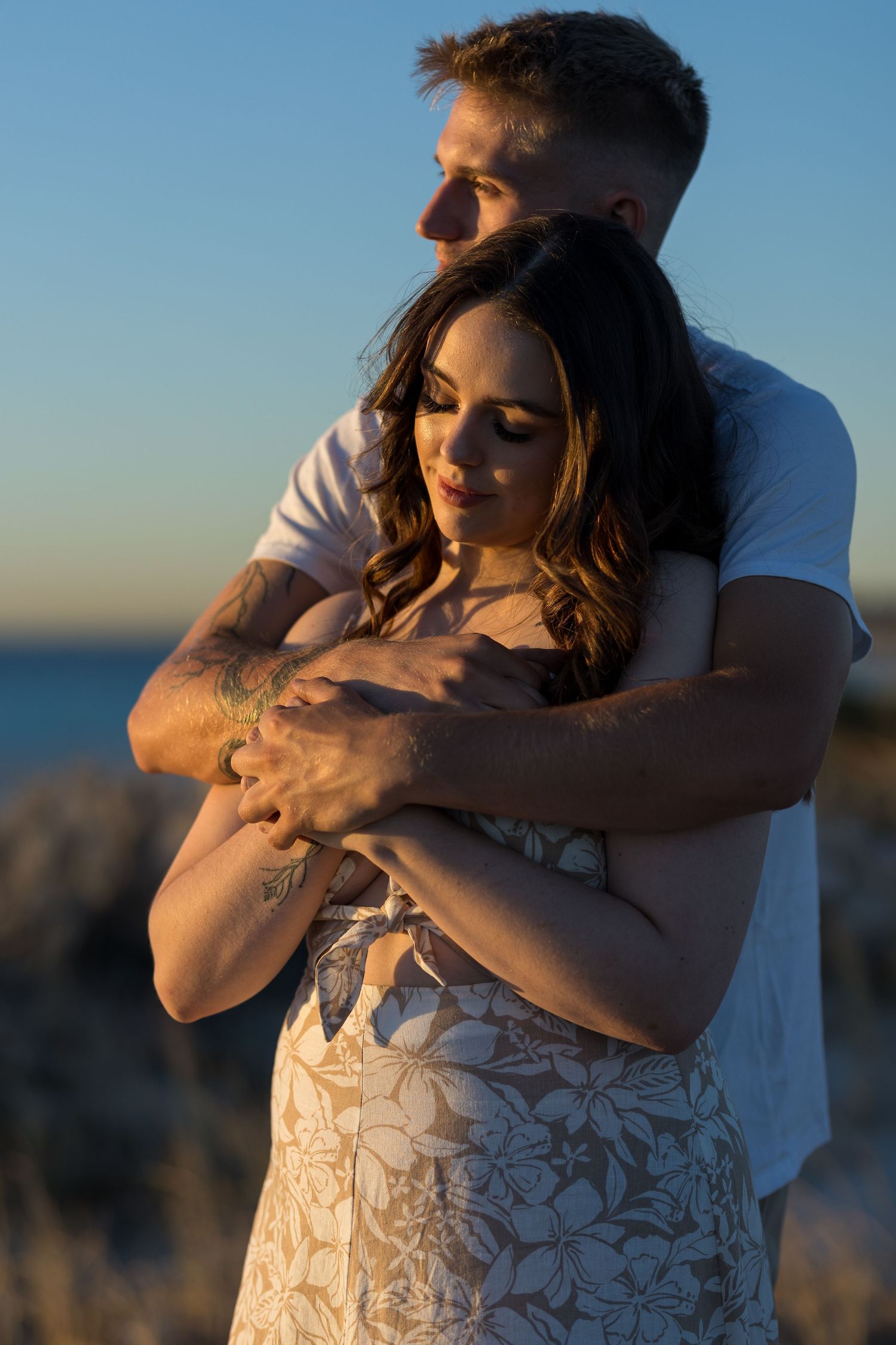 Man embracing woman from behind at sunset near rocky coastline.