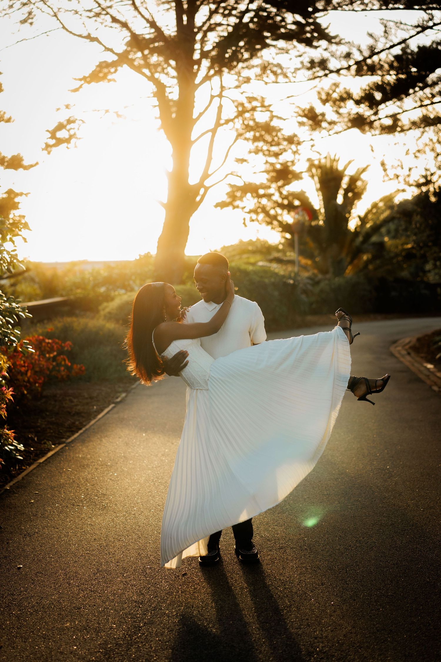 Man in white shirt carries a woman in a flowing white pleated dress along a tree-lined path at golden hour sunset.