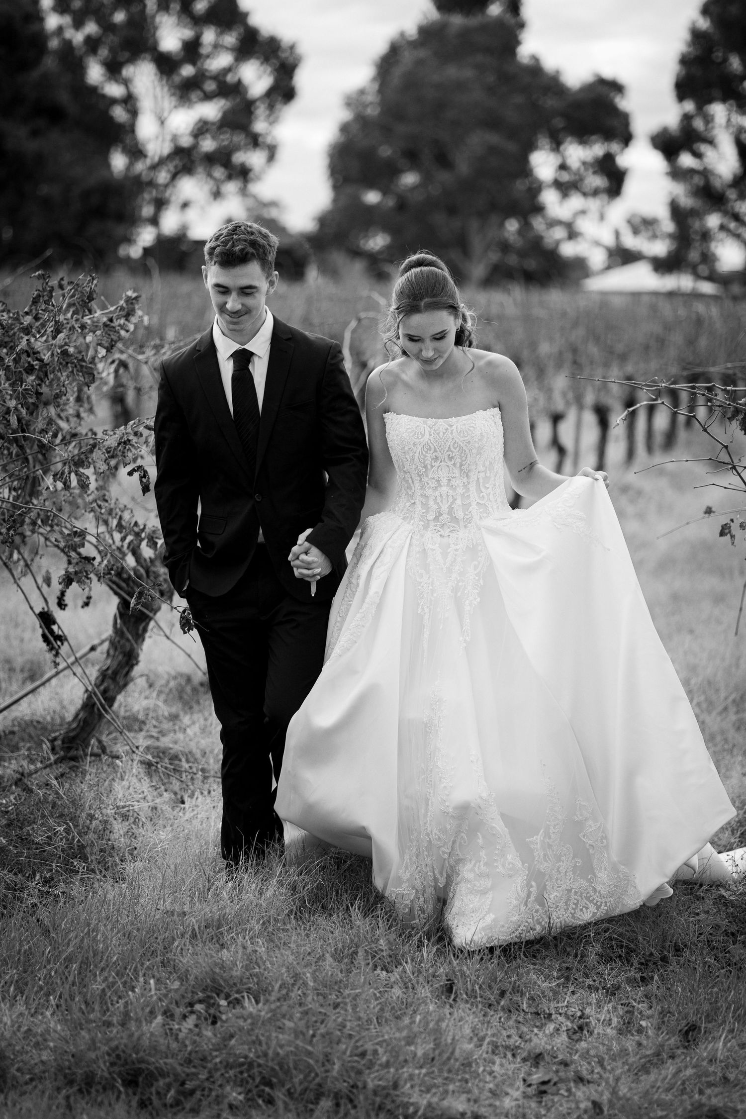 Bride and groom walk hand in hand through vineyard in black and white wedding portrait.