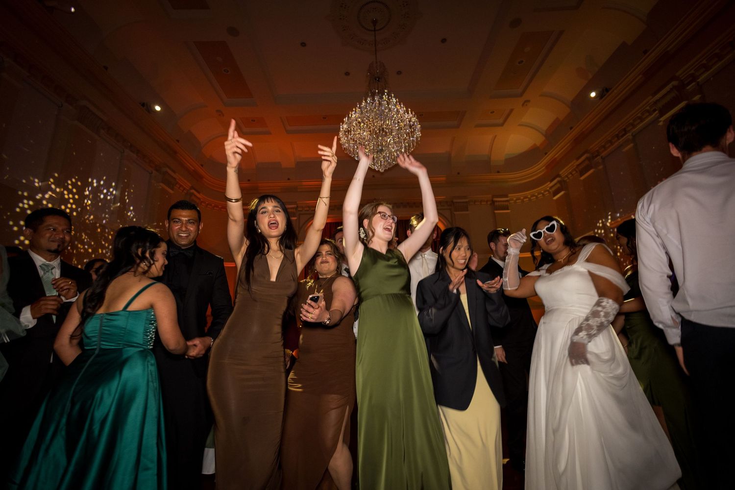 Wedding guests dance energetically on ballroom floor reaching toward ornate chandelier at reception at Montgomery Hall
