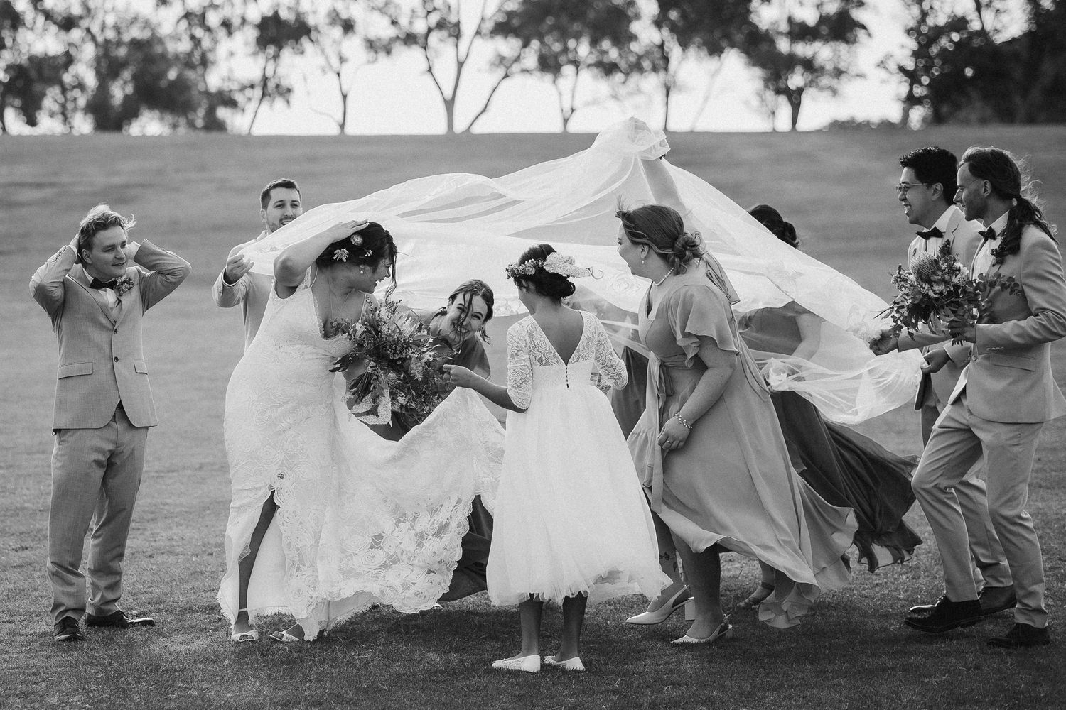 Black and white photo of wedding party laughing as the bride's veil billows over the group outdoors.