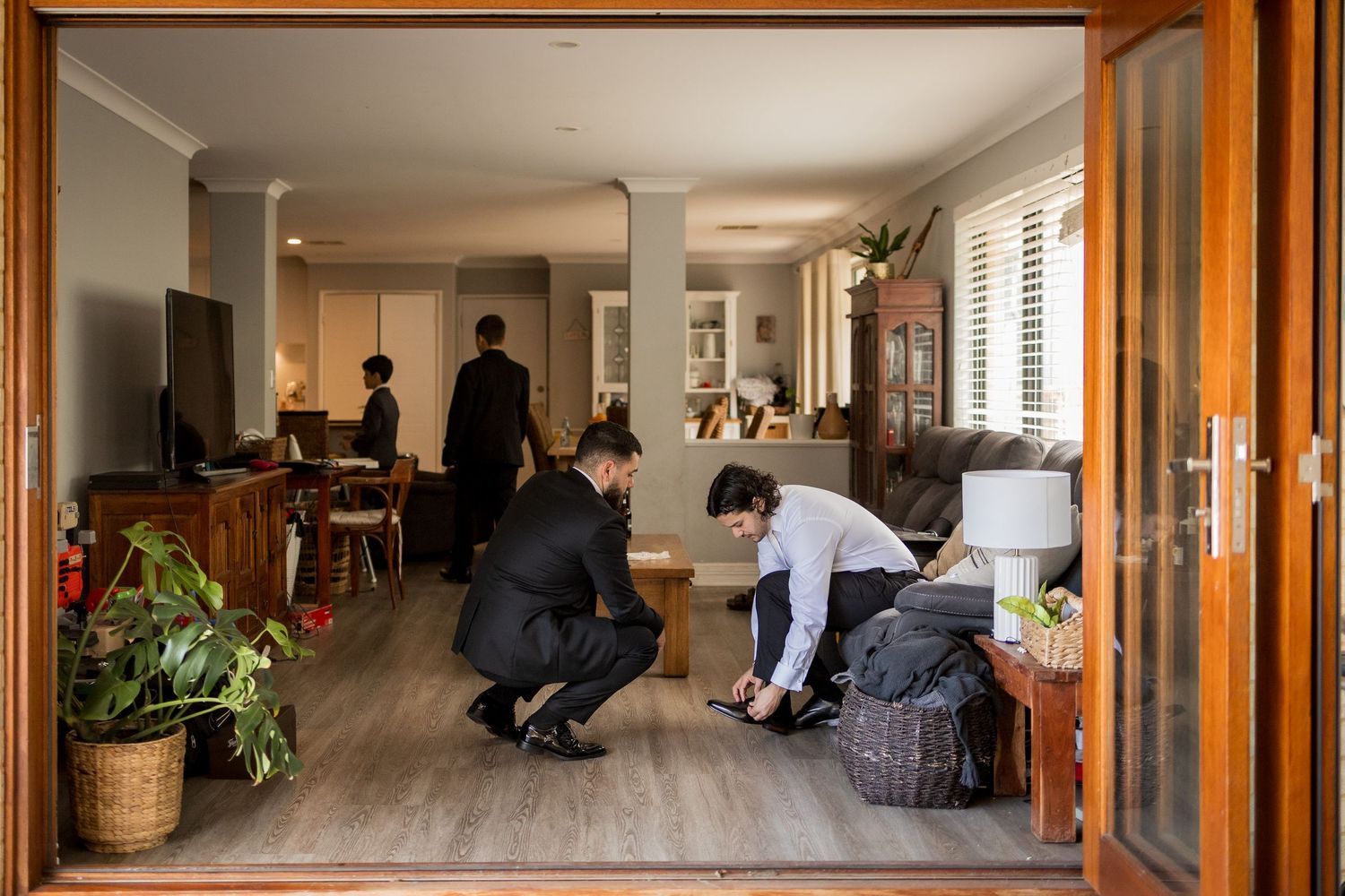 Men in formal black suits preparing in a stylish living room, one crouching to adjust shoes before a wedding.
