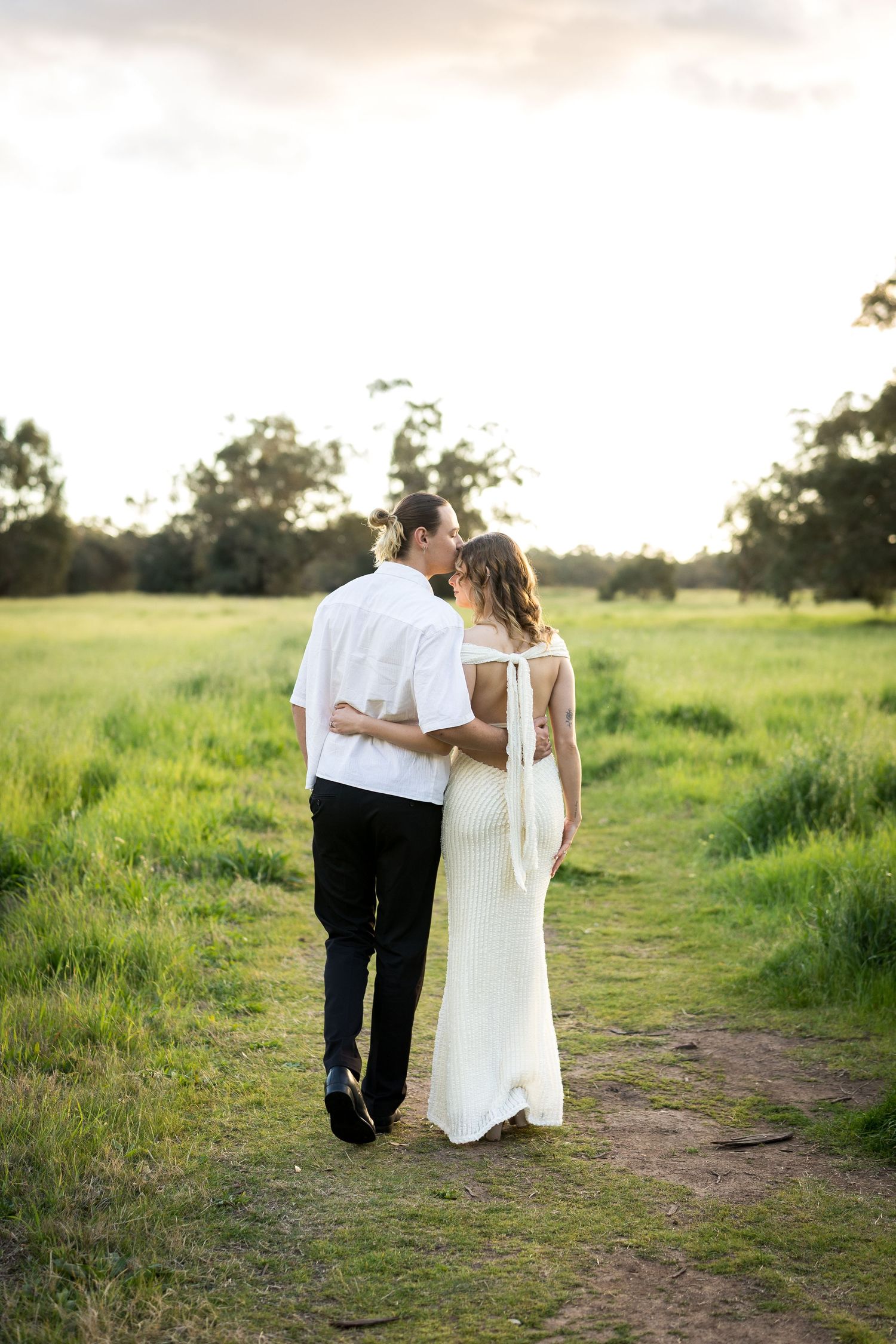 Couple kissing while walking through a lush green field at sunset, bride in white gown and groom in white shirt.