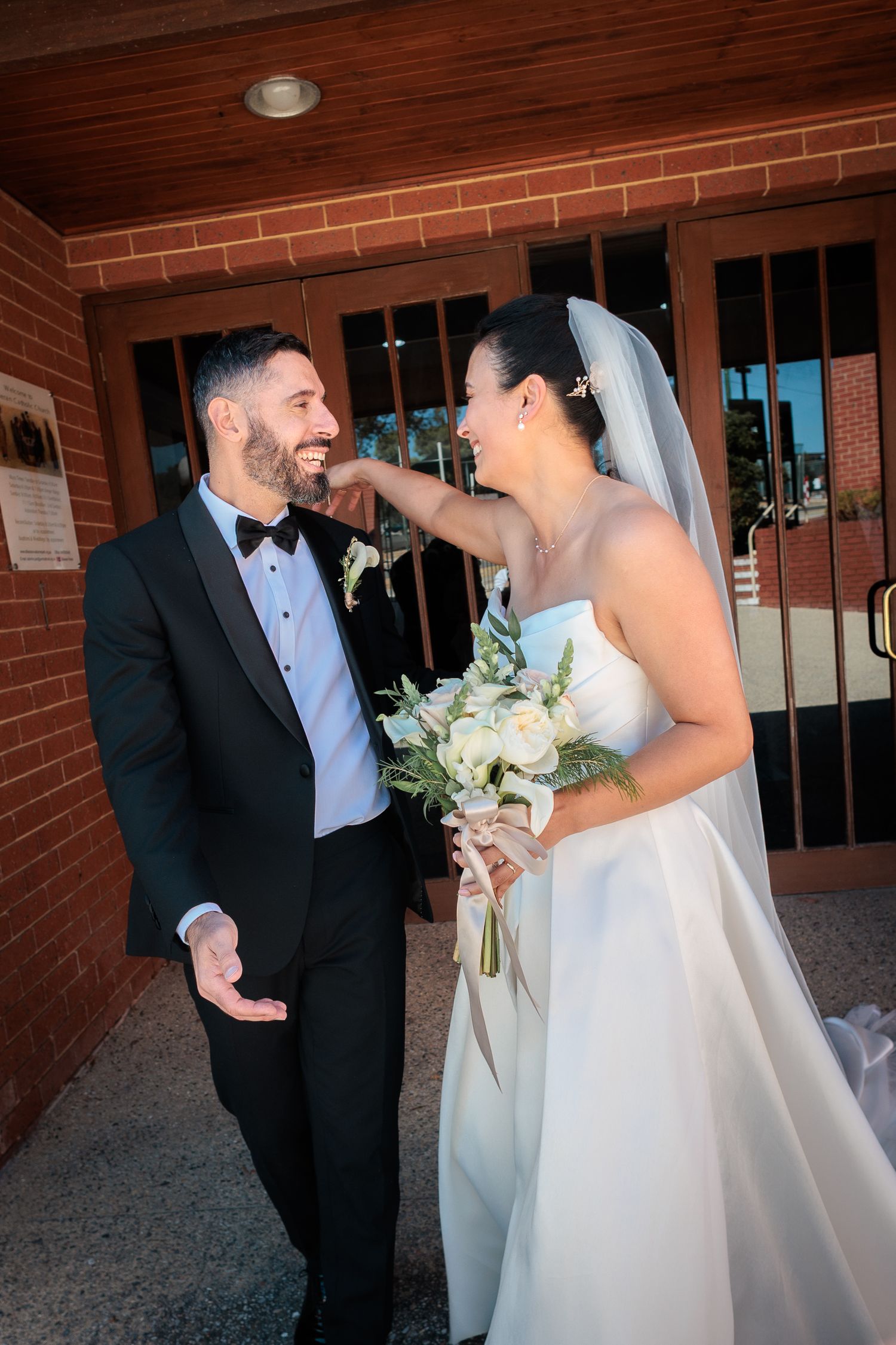 Bride in white gown holding bouquet laughs with groom in tuxedo outside brick church on wedding day.