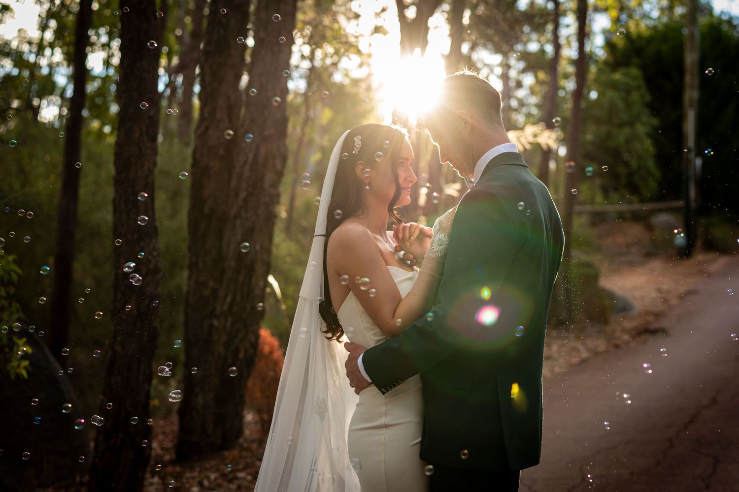 Bride and groom share a romantic moment in sunlit forest with bubbles, bride in white gown with veil.