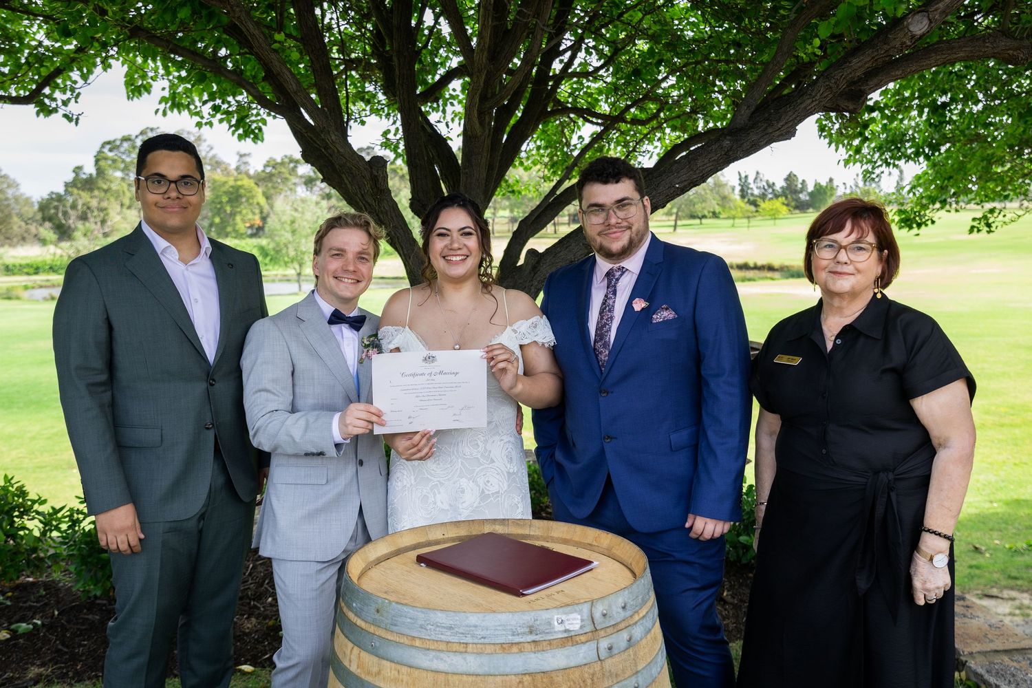Bride and groom hold marriage certificate beside wine barrel with three guests under tree at wedding venue.
