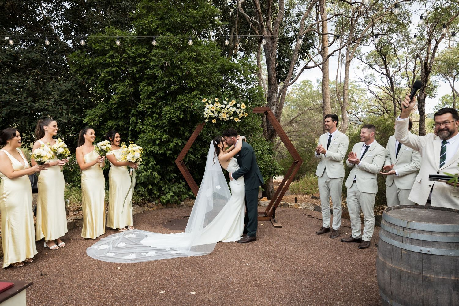 Bride and groom share first kiss at outdoor wedding ceremony with bridesmaids in yellow dresses and groomsmen cheering.